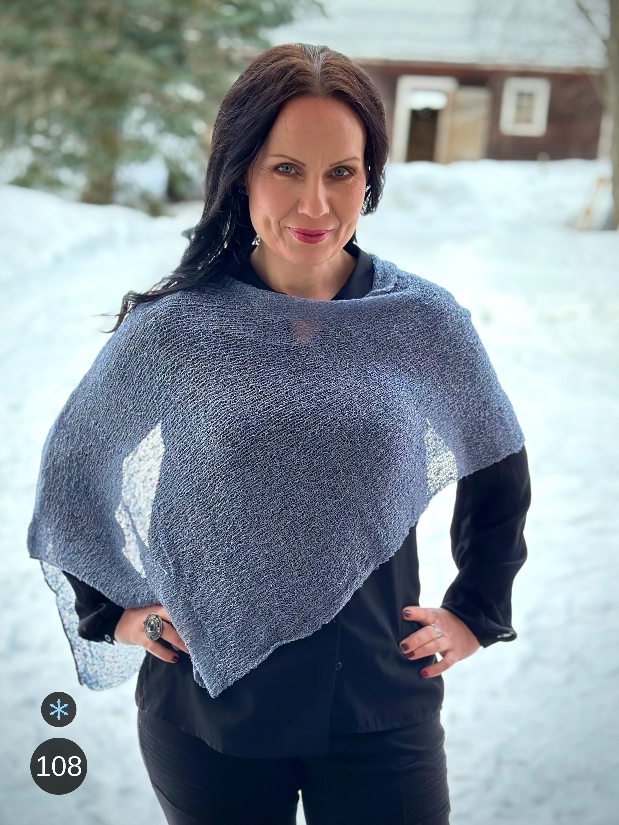 Kvinna bär Ella Cape i färg 108 Granit, en kall färg i granitgrå med sval blå underton, finstickad cape som draperas mjukt över axlarna, fotad mot snön framför en röd byggnad. Ulla Jacobsson Kläder.