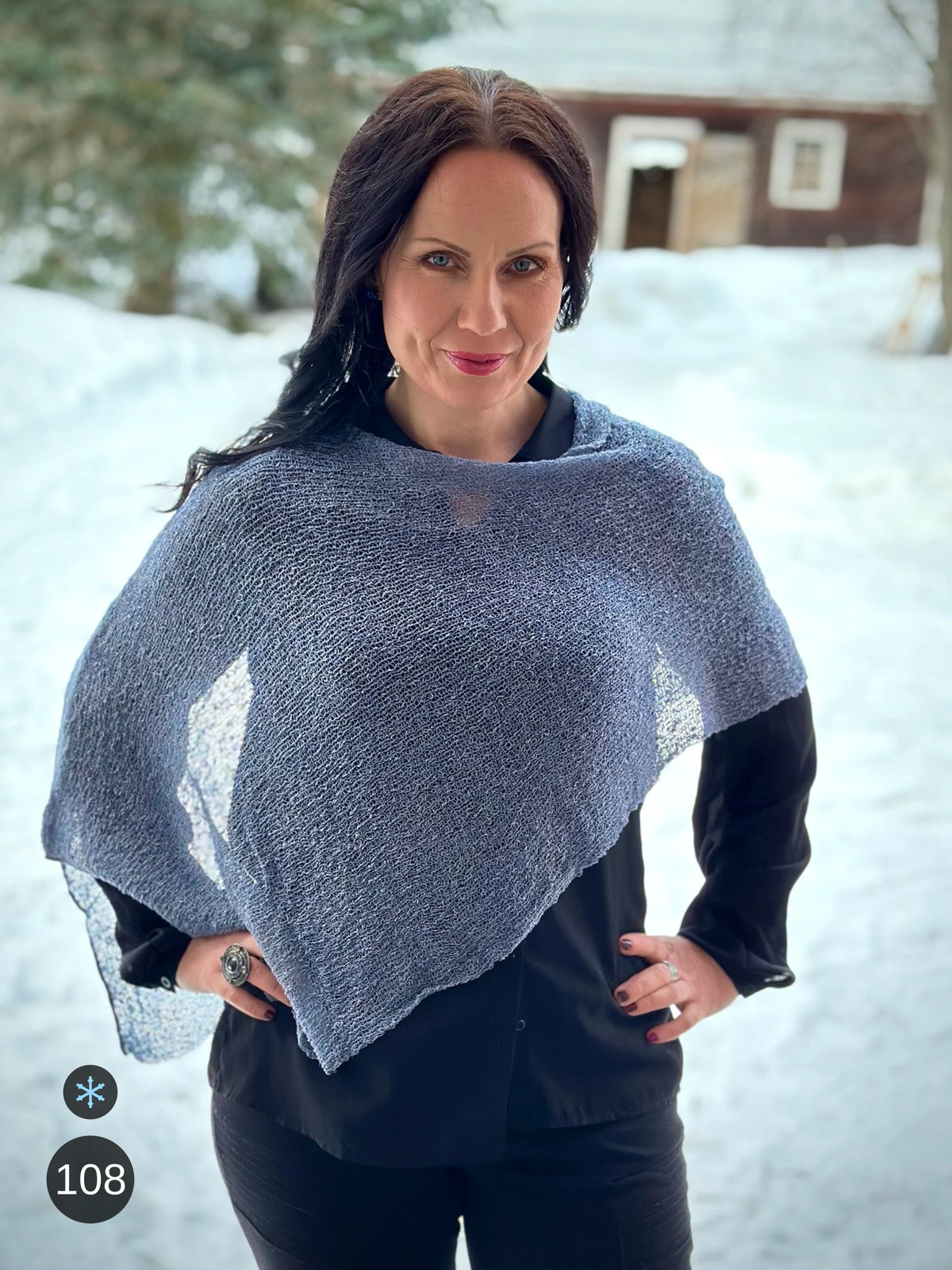 Kvinna bär Ella Cape i färg 108 Stålblå, en kall färg i stålblågrå med sval blå underton, finstickad cape som draperas mjukt över axlarna, fotad mot snön framför en röd byggnad. Ulla Jacobsson Kläder.