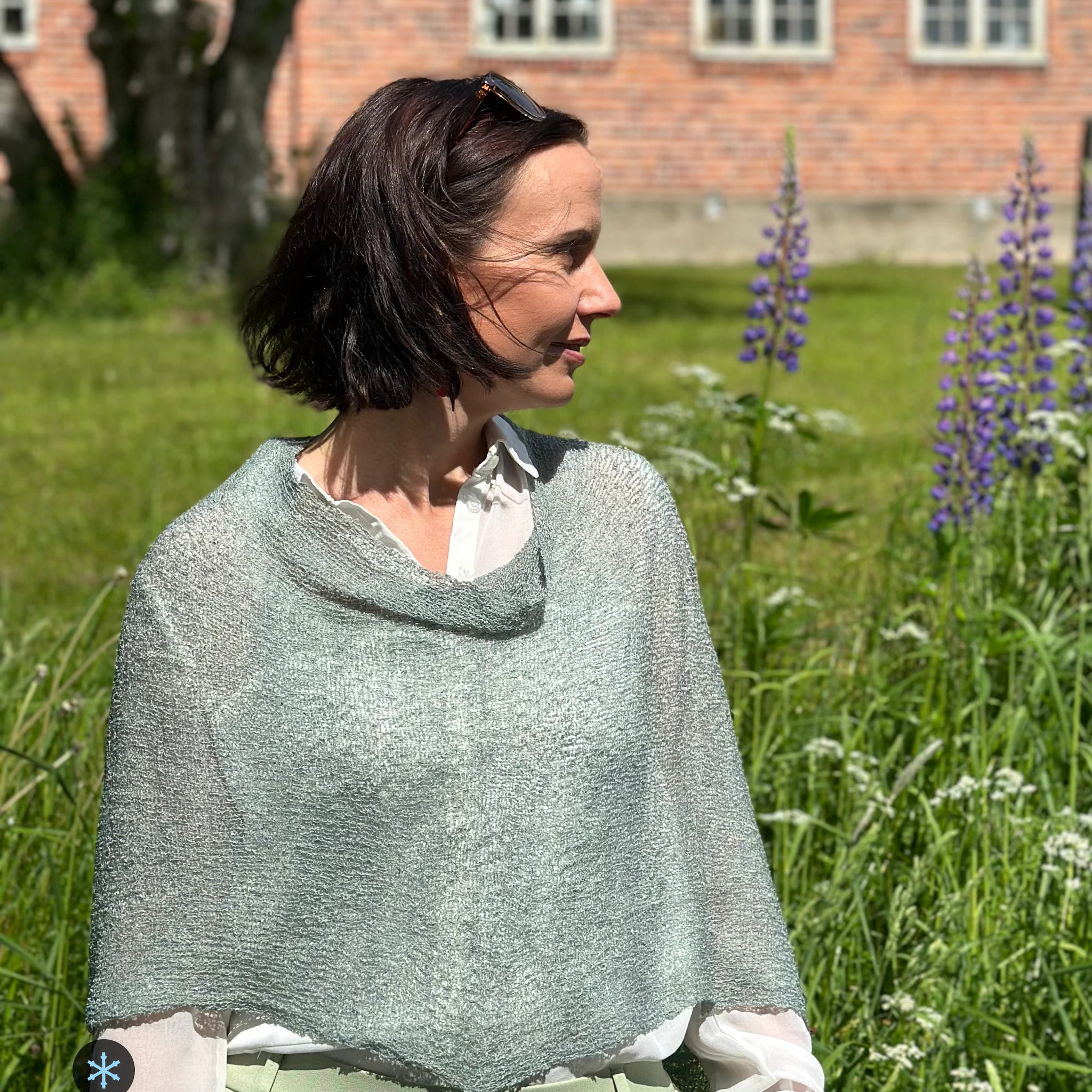 Kvinna bär Ella Cape i färg 109 Grå mossa, en kall färg i dämpad gråmossgrön, finstickad cape som draperas mjukt över axlarna, fotad i en sommarträdgård med blommande lupiner. Ulla Jacobsson Kläder.