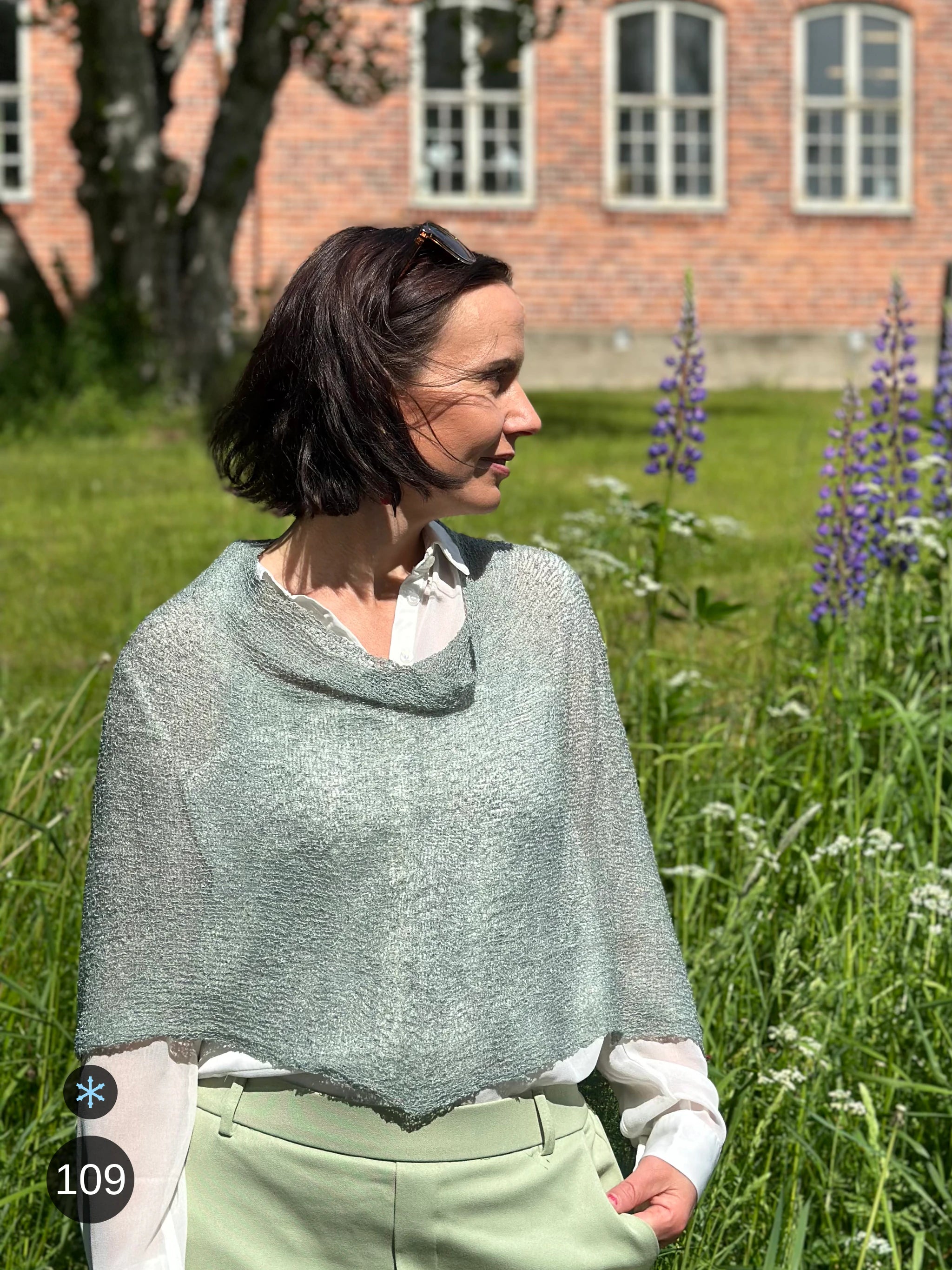 Kvinna bär Ella Cape i färg 109 Grå mossa, en kall färg i dämpad gråmossgrön, finstickad cape som draperas mjukt över axlarna, fotad i en sommarträdgård med blommande lupiner. Ulla Jacobsson Kläder.