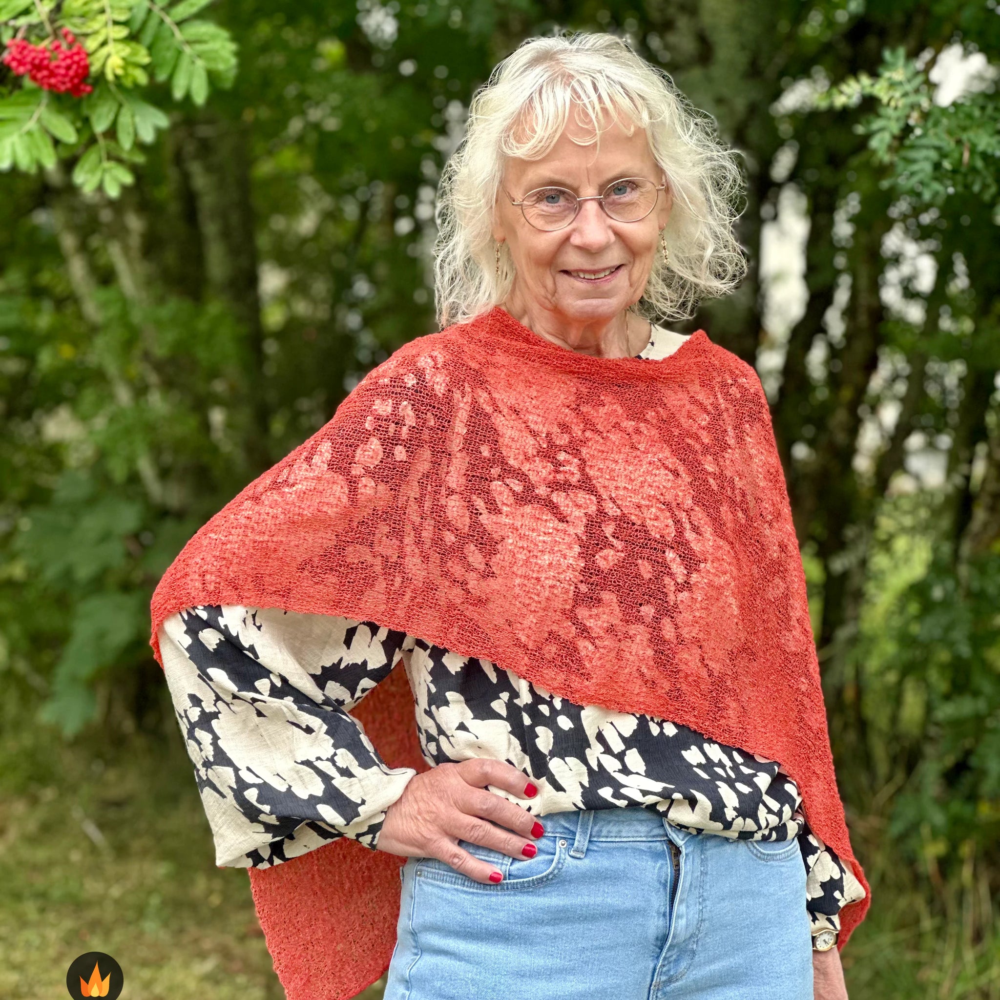 Kvinna bär Ella Cape i färg 111 Rost, en varm färg i varm rostorange, finstickad cape som draperas mjukt över axlarna, fotad utomhus i grönska. Ulla Jacobsson Kläder.