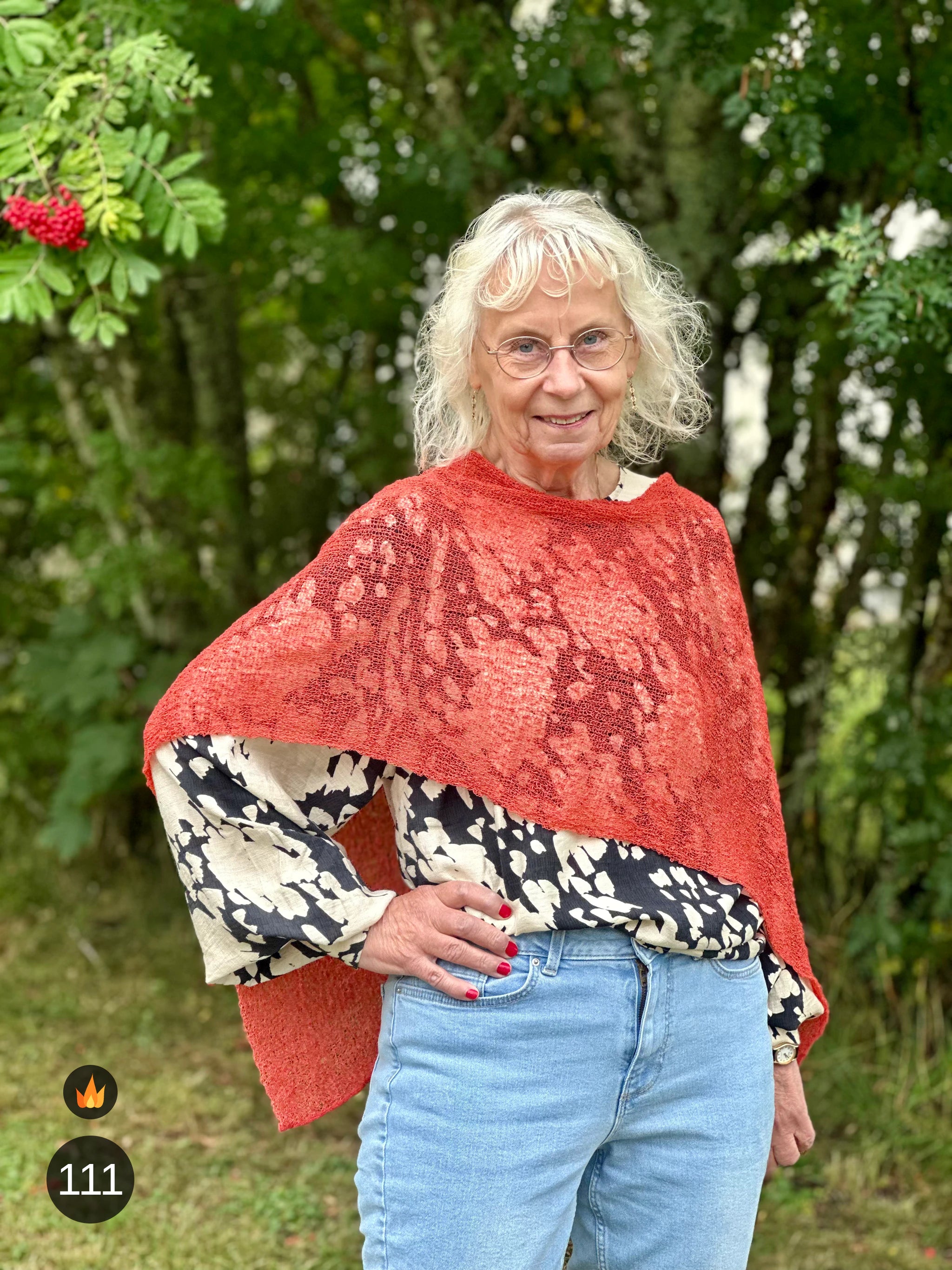 Kvinna bär Ella Cape i färg 111 Rost, en varm färg i varm rostorange, finstickad cape som draperas mjukt över axlarna, fotad utomhus i grönska. Ulla Jacobsson Kläder.