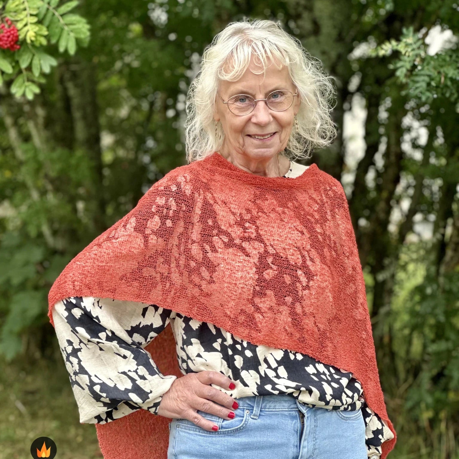 Kvinna bär Ella Cape i färg 111 Rost, en varm färg, finstickad cape som draperas mjukt över axlarna, fotad utomhus i grönska. Ulla Jacobsson Kläder.