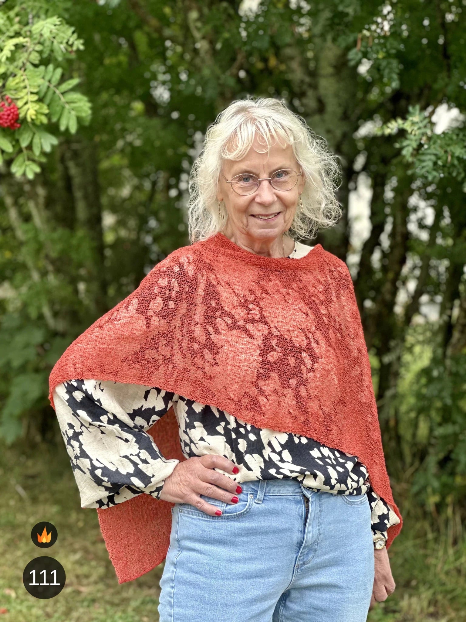 Kvinna bär Ella Cape i färg 111 Rost, en varm färg, finstickad cape som draperas mjukt över axlarna, fotad utomhus i grönska. Ulla Jacobsson Kläder.