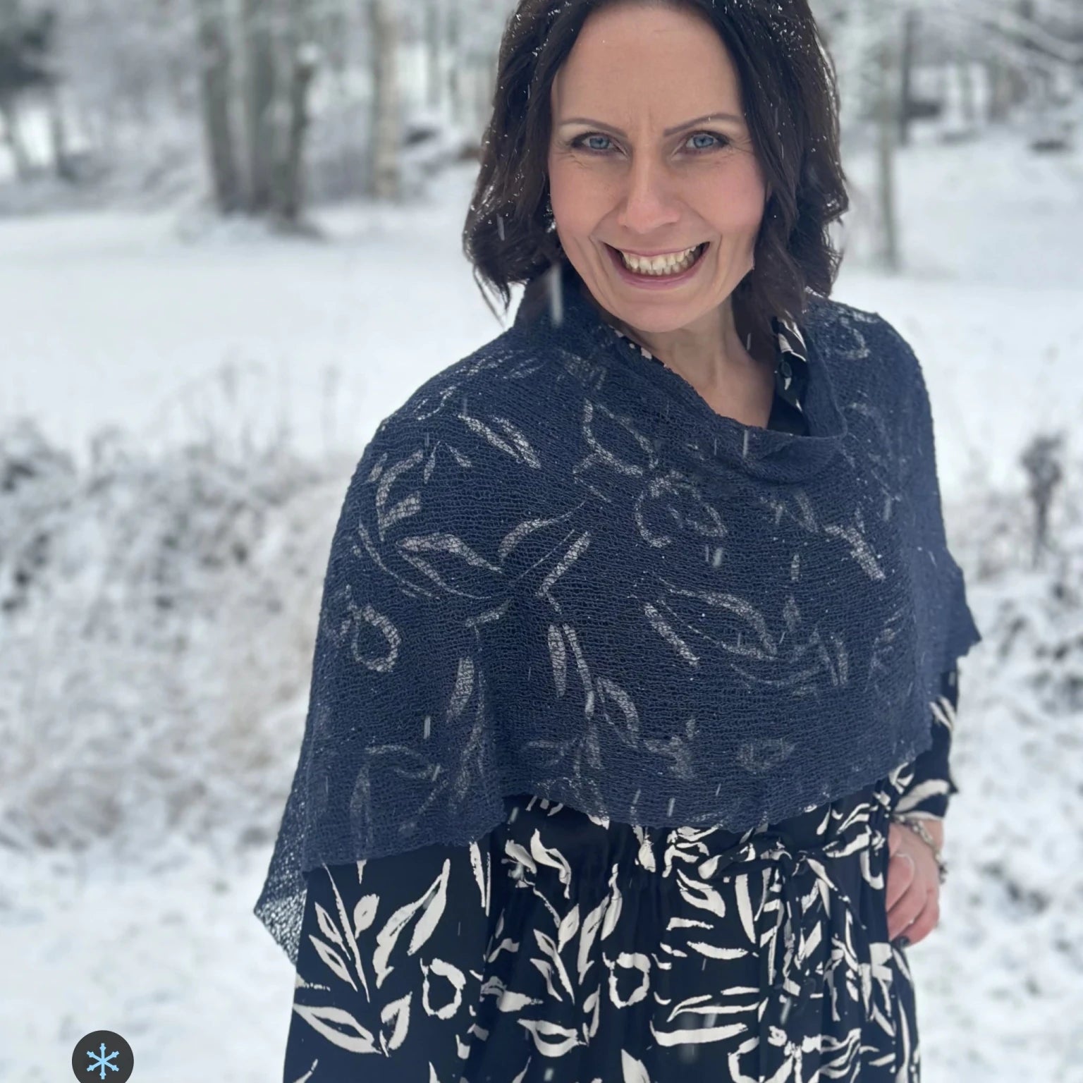 Kvinna bär Ella Cape i färg 112 Torr marin, en kall färg, finstickad cape som draperas mjukt över axlarna, fotad i ett snöigt vinterlandskap. Ulla Jacobsson Kläder.