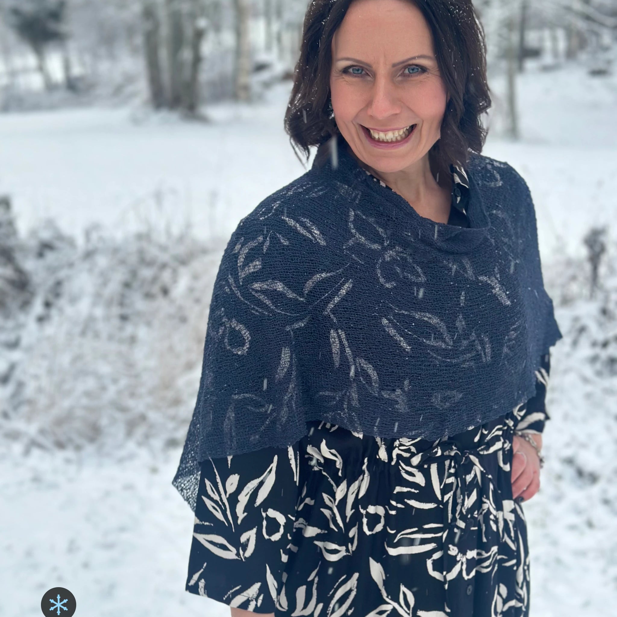 Kvinna bär Ella Cape i färg 112 Torr marin, en kall färg, finstickad cape som draperas mjukt över axlarna, fotad i ett snöigt vinterlandskap. Ulla Jacobsson Kläder.