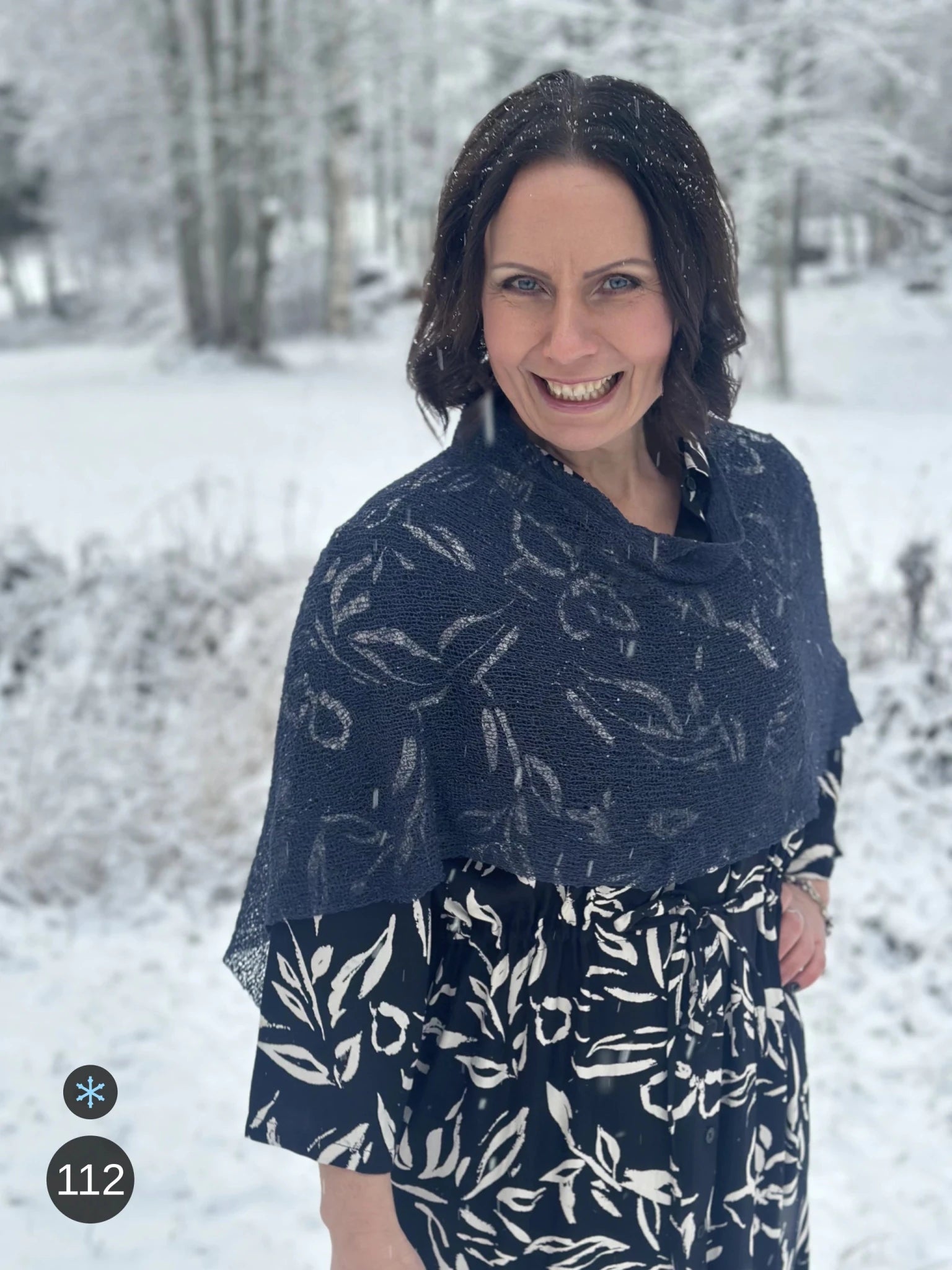Kvinna bär Ella Cape i färg 112 Torr marin, en kall färg, finstickad cape som draperas mjukt över axlarna, fotad i ett snöigt vinterlandskap. Ulla Jacobsson Kläder.