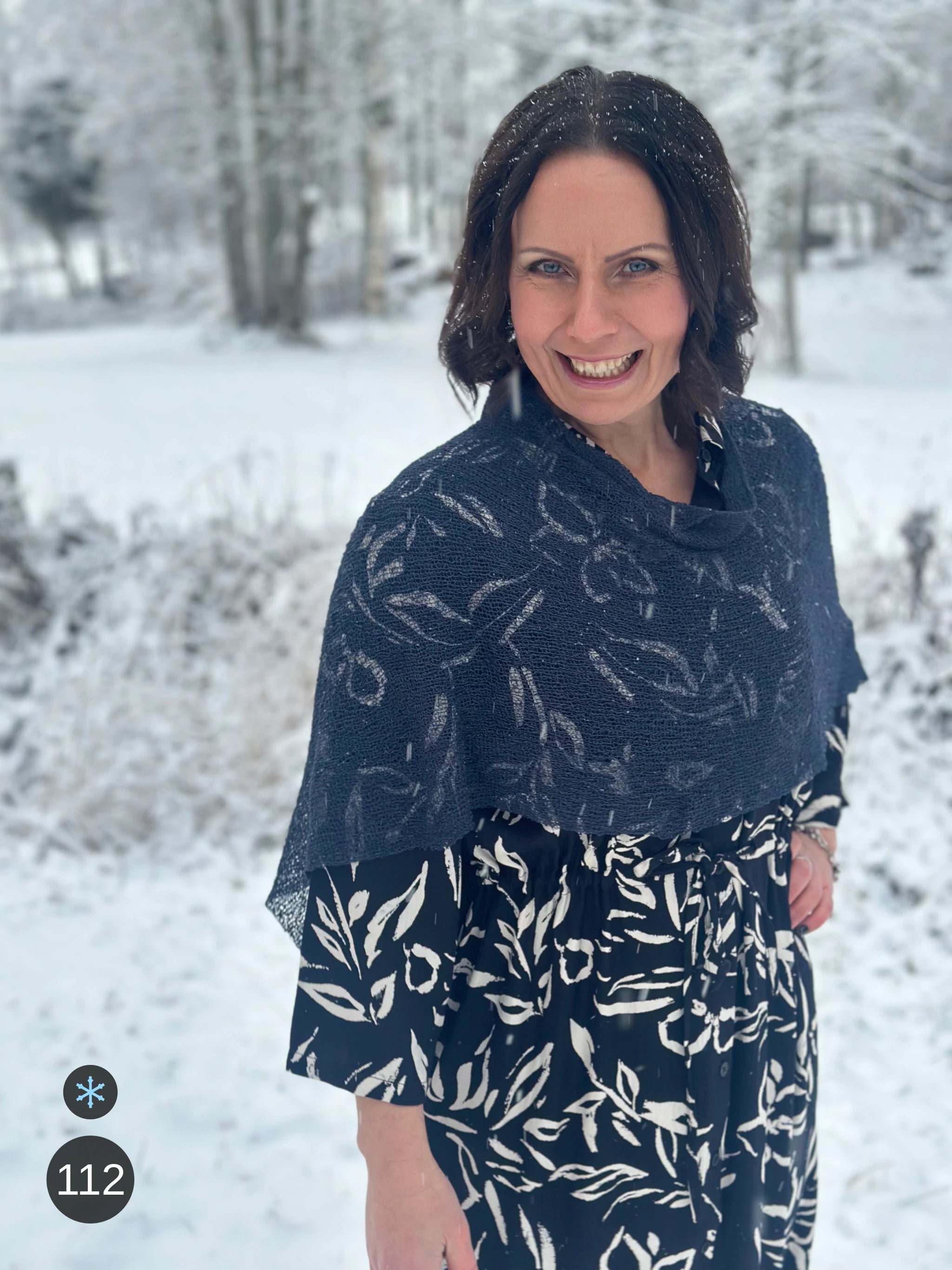 Kvinna bär Ella Cape i färg 112 Torr marin, en kall färg, finstickad cape som draperas mjukt över axlarna, fotad i ett snöigt vinterlandskap. Ulla Jacobsson Kläder.