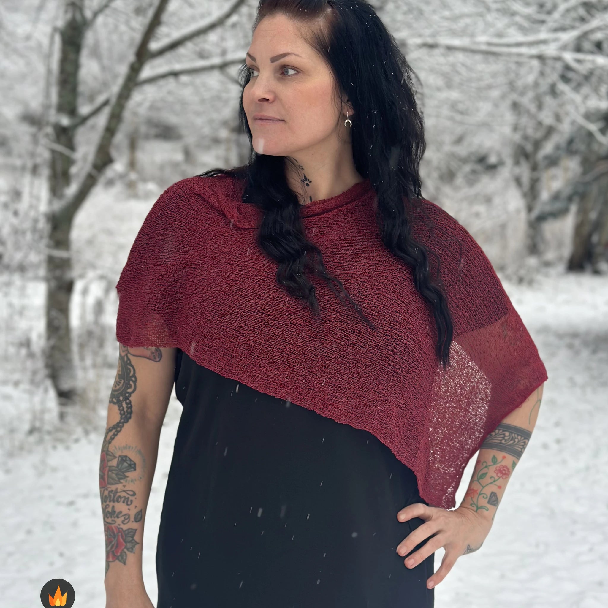 Kvinna bär Ella Cape i färg 114 Maroon, en varm färg, finstickad cape som draperas mjukt över axlarna, fotad i vintrigt snölandskap. Ulla Jacobsson Kläder.
