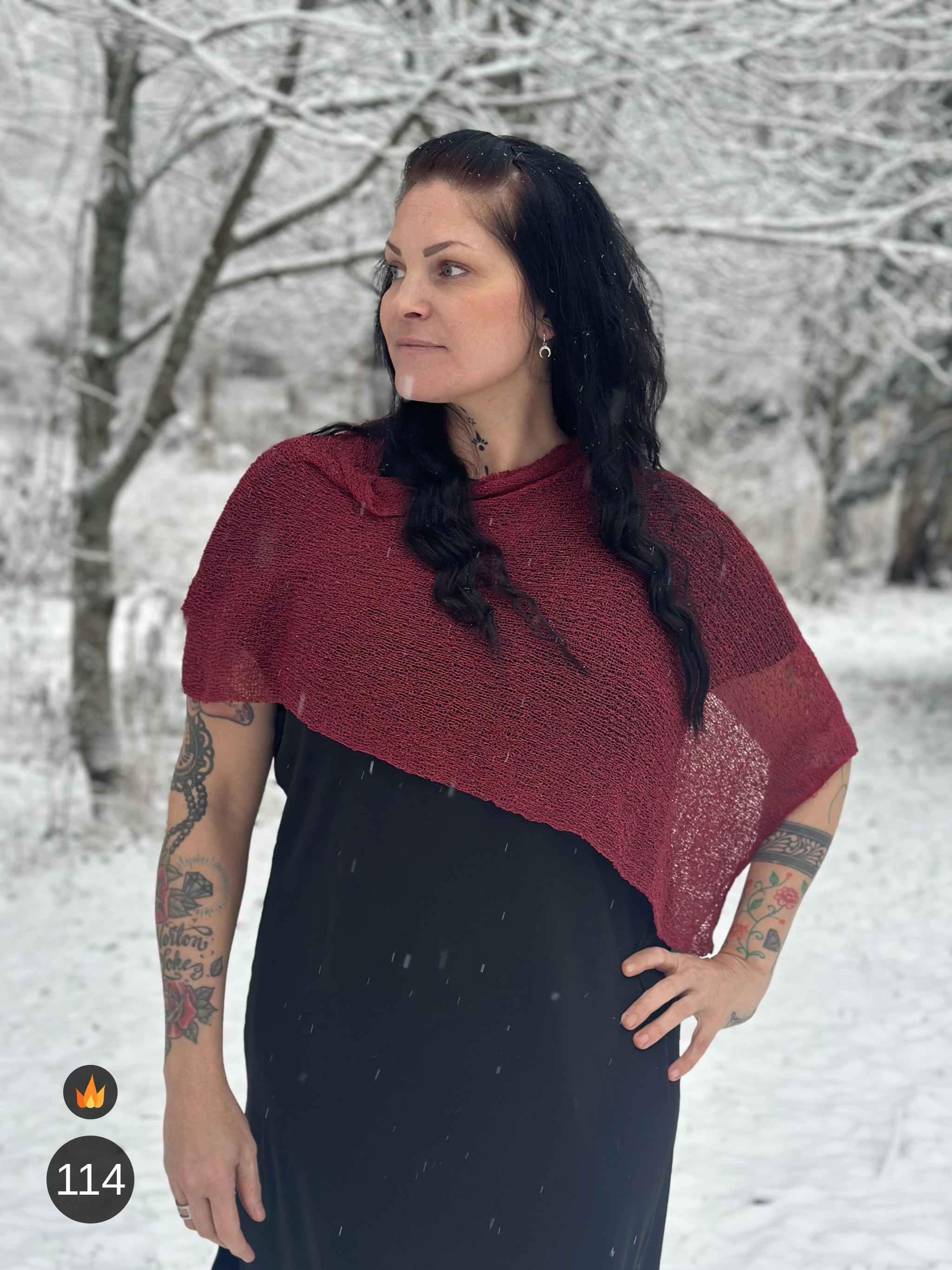 Kvinna bär Ella Cape i färg 114 Maroon, en varm färg, finstickad cape som draperas mjukt över axlarna, fotad i vintrigt snölandskap. Ulla Jacobsson Kläder.