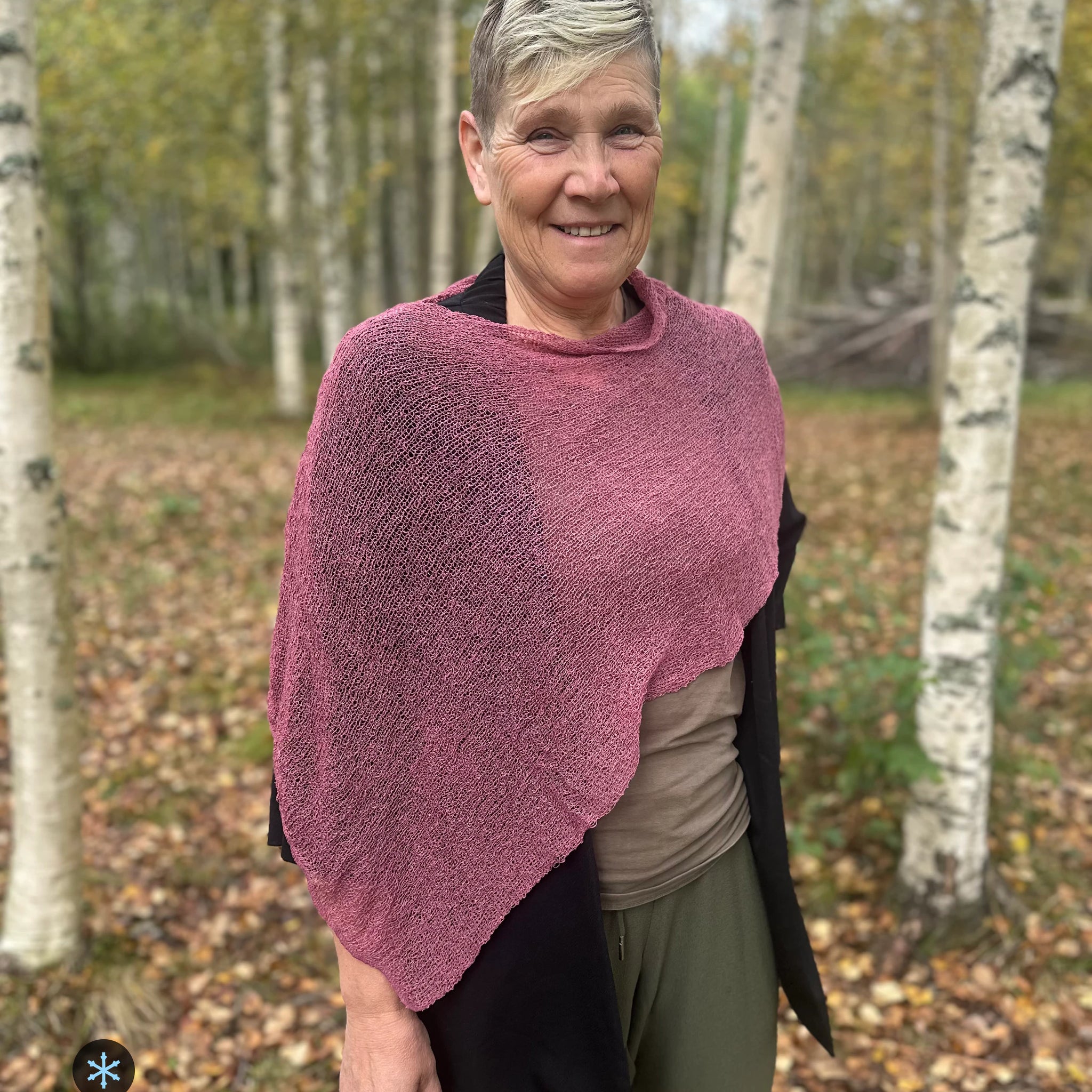 Kvinna bär Ella Cape i färg 115 Ljung, en kall färg i ljungrosa med sval underton, finstickad cape som draperas mjukt över axlarna, fotad bland höstgula björkar. Ulla Jacobsson Kläder.