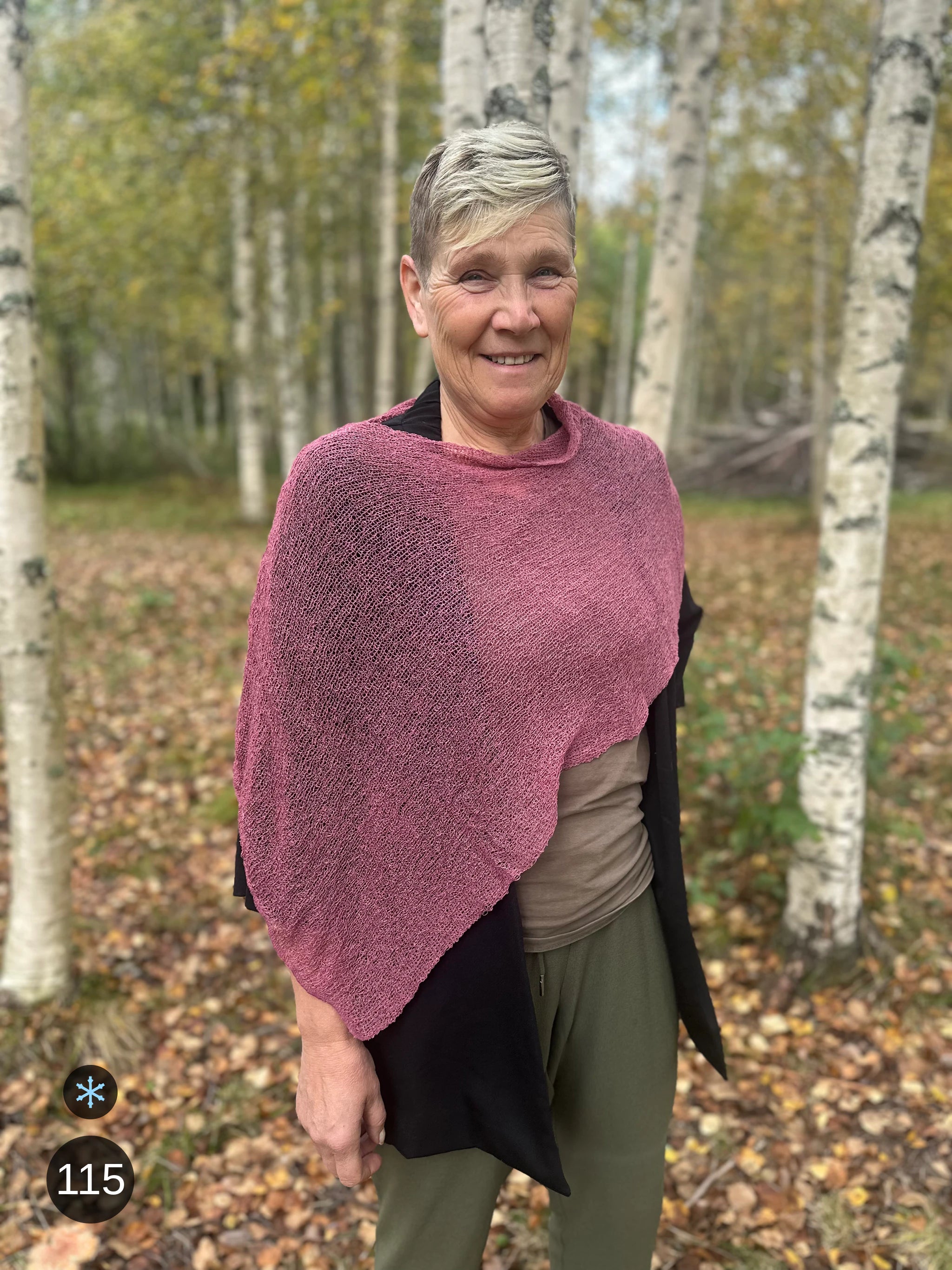 Kvinna bär Ella Cape i färg 115 Ljung, en kall färg i ljungrosa med sval underton, finstickad cape som draperas mjukt över axlarna, fotad bland höstgula björkar. Ulla Jacobsson Kläder.