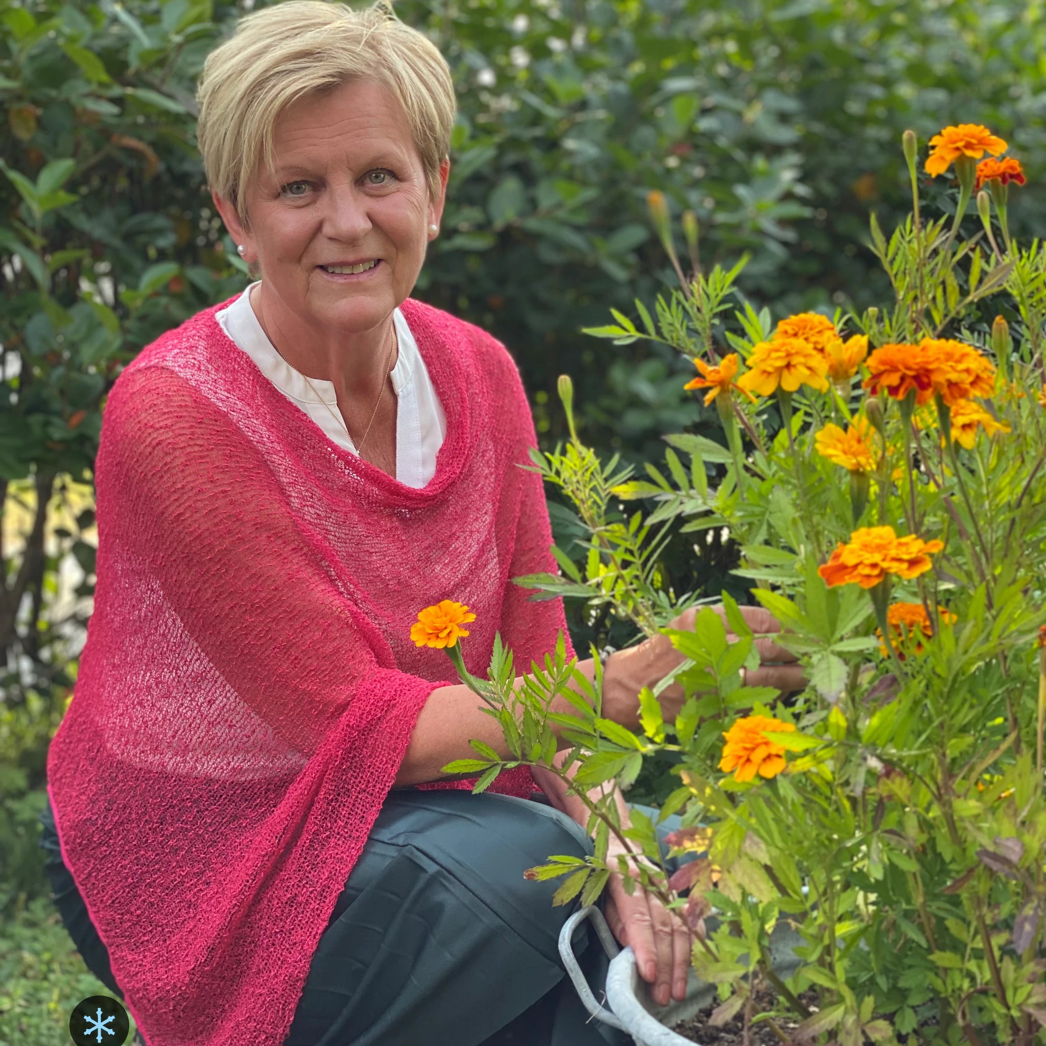Kvinna bär Ella Cape i färg 116 Azaleröd, en kall färg i azaleröd med sval blå underton, finstickad cape som draperas mjukt över axlarna, fotad vid en zinkbalja med orange ringblommor. Ulla Jacobsson Kläder.