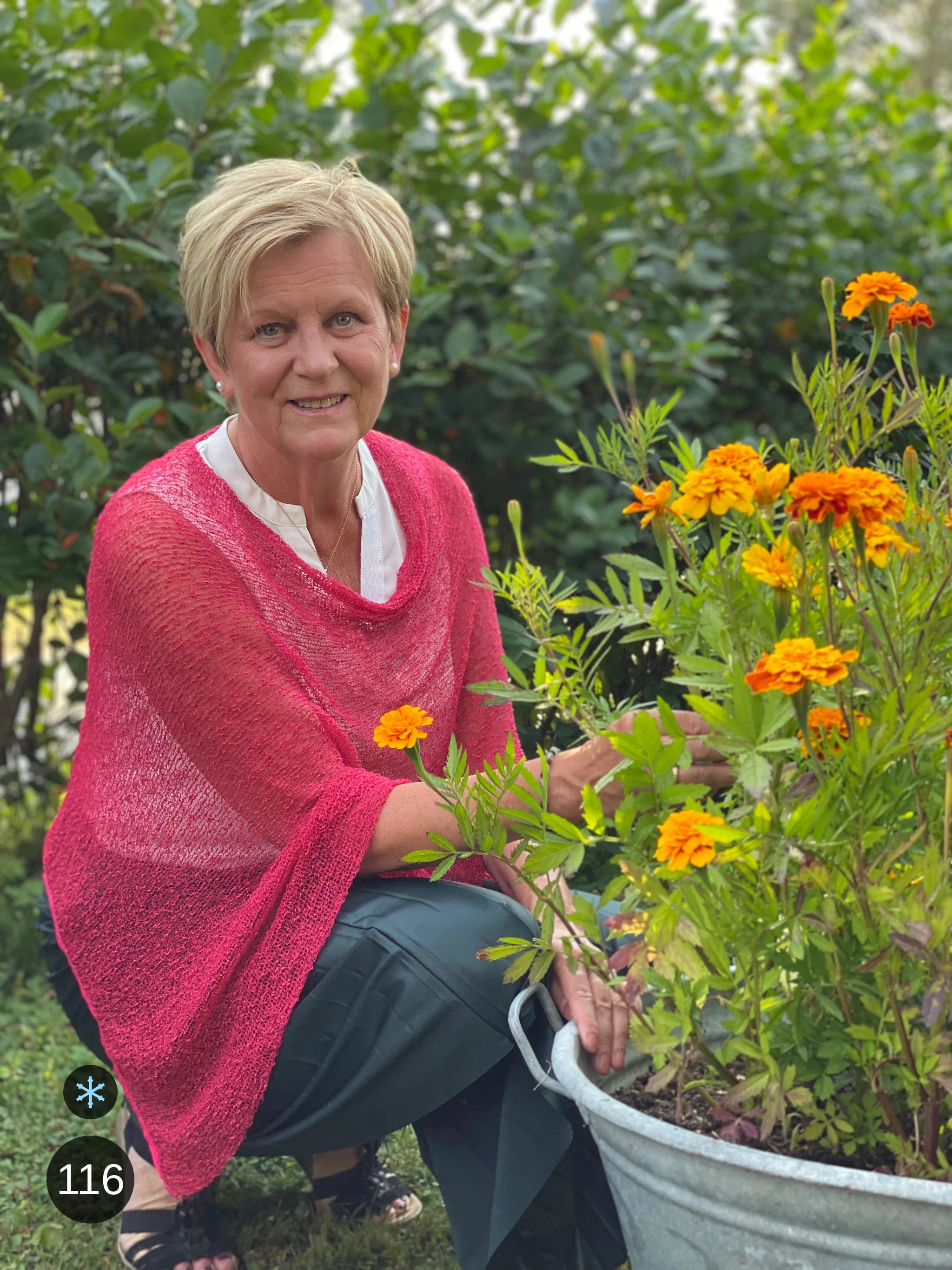 Kvinna bär Ella Cape i färg 116 Azaleröd, en kall färg i azaleröd med sval blå underton, finstickad cape som draperas mjukt över axlarna, fotad vid en zinkbalja med orange ringblommor. Ulla Jacobsson Kläder.