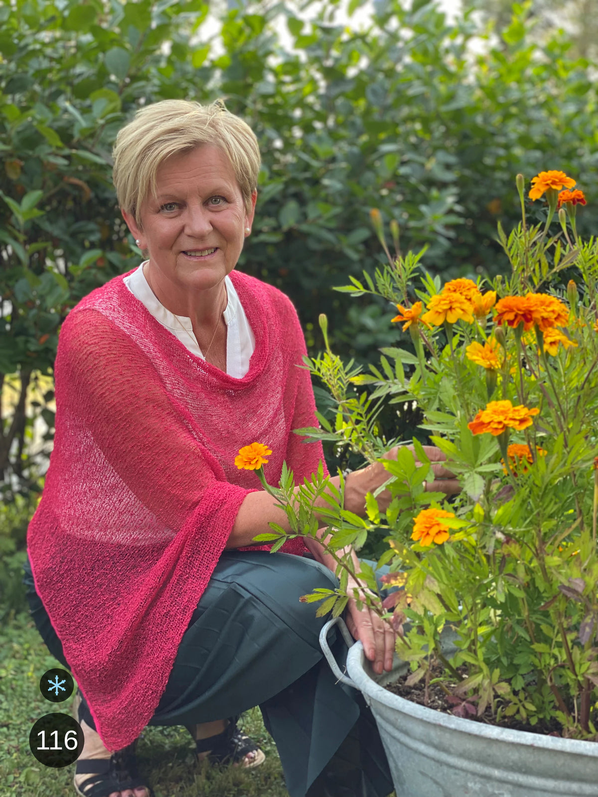 Kvinna bär Ella Cape i färg 116 Hallonrosa, en kall färg i hallonrosa med sval blå underton, finstickad cape som draperas mjukt över axlarna, fotad vid en zinkbalja med orange ringblommor. Ulla Jacobsson Kläder.