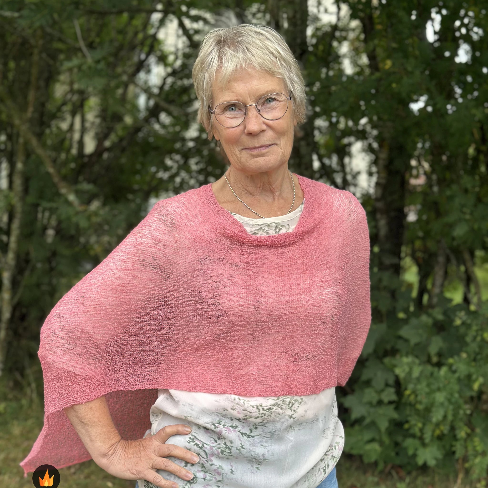Kvinna bär Ella Cape i färg 118 Gammelrosa, en varm färg, finstickad cape som draperas mjukt över axlarna, fotad utomhus i grönska. Ulla Jacobsson Kläder.