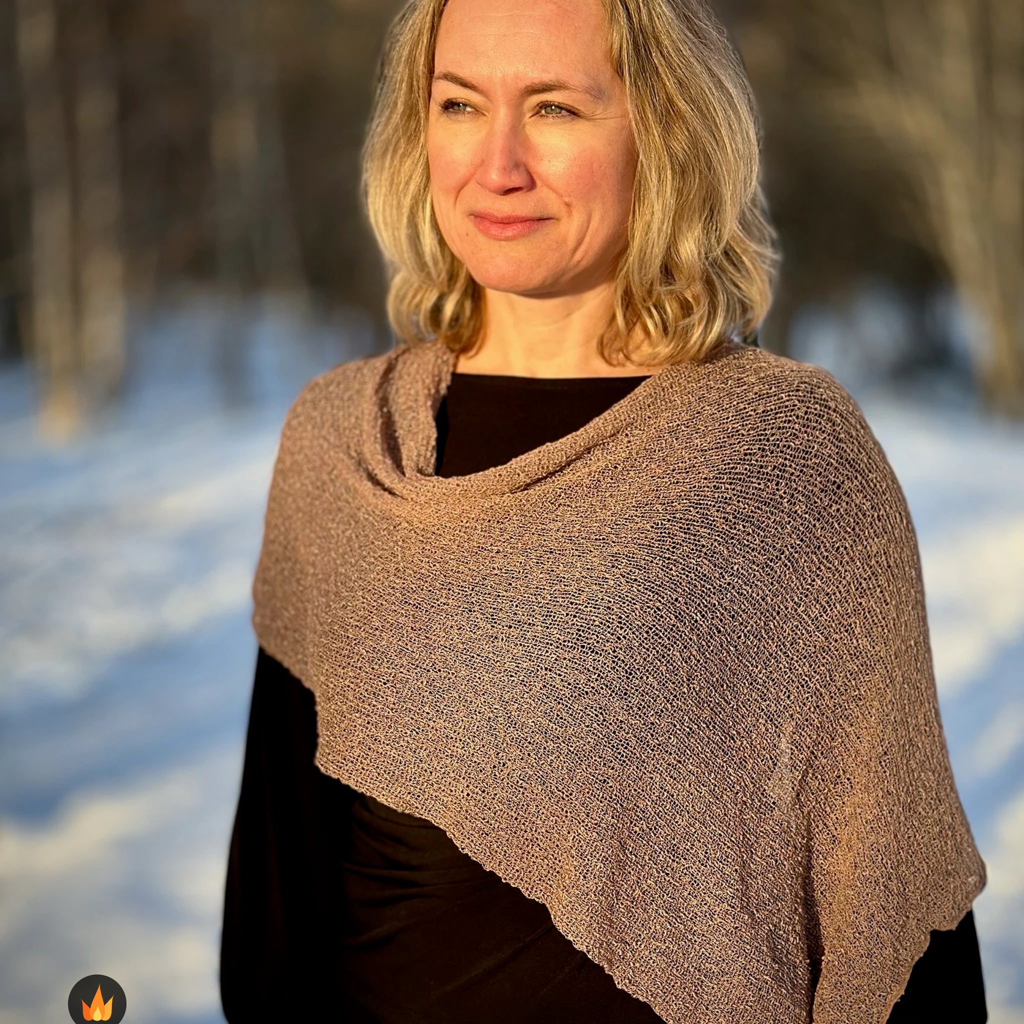 Kvinna bär Ella Cape i färg 119 Cashmere, en varm färg i cashmerebeige med gyllenbrun värme, finstickad cape som draperas mjukt över axlarna, fotad på en snöig väg. Ulla Jacobsson Kläder.
