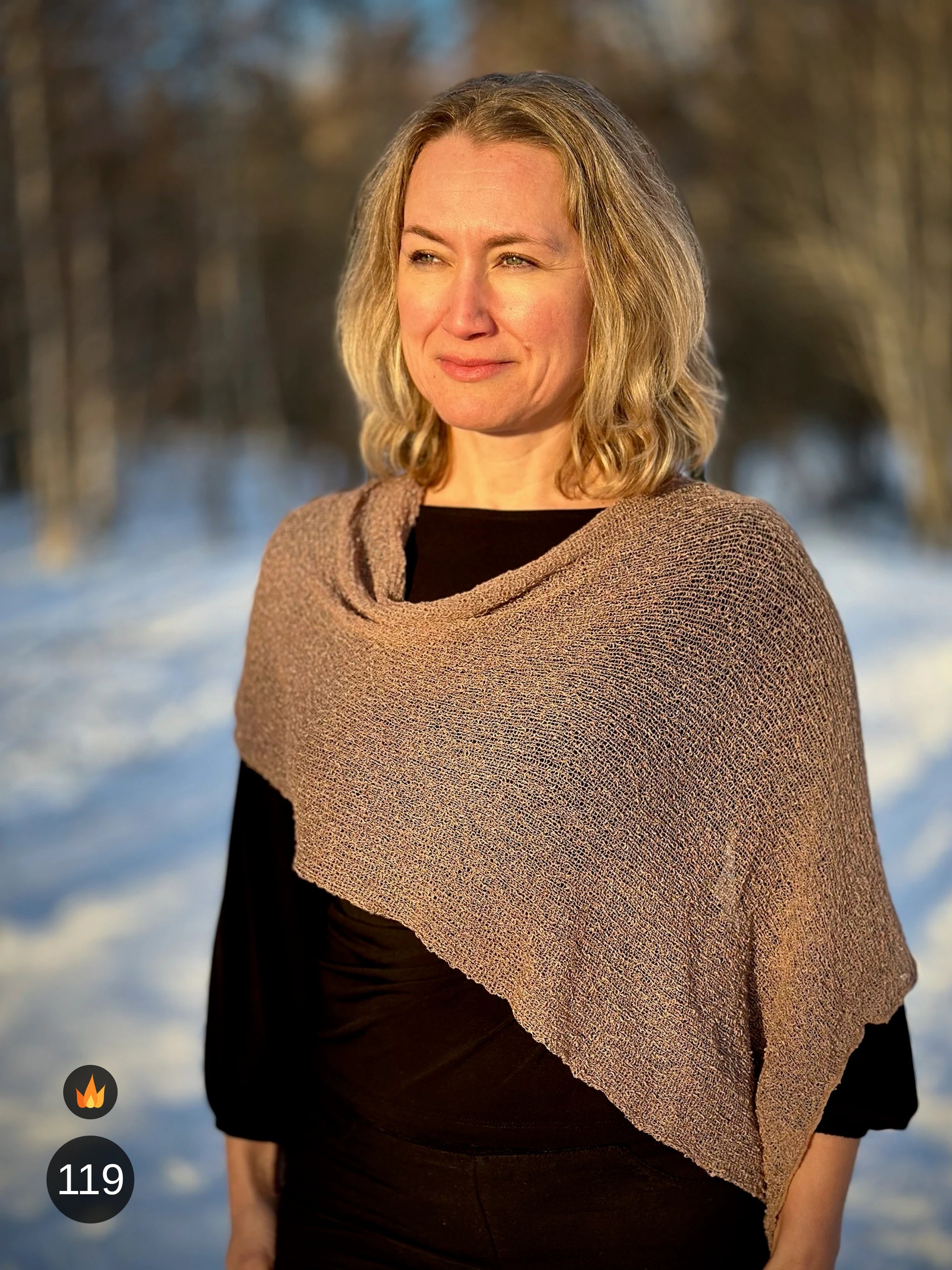 Kvinna bär Ella Cape i färg 119 Cashmere, en varm färg i cashmerebeige med gyllenbrun värme, finstickad cape som draperas mjukt över axlarna, fotad på en snöig väg. Ulla Jacobsson Kläder.