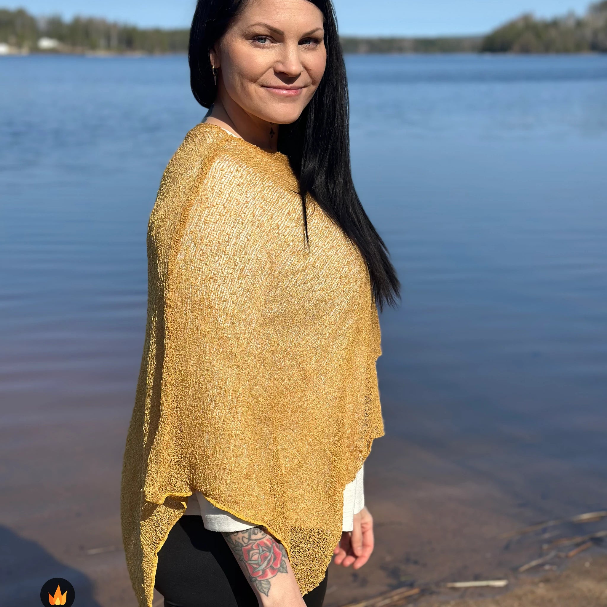 Kvinna bär Ella Cape i färg 121 Ljus senap, en varm färg i ljus senapsgul med gyllene skimmer, finstickad cape som draperas mjukt över axlarna, fotad vid en stilla sjö. Ulla Jacobsson Kläder.