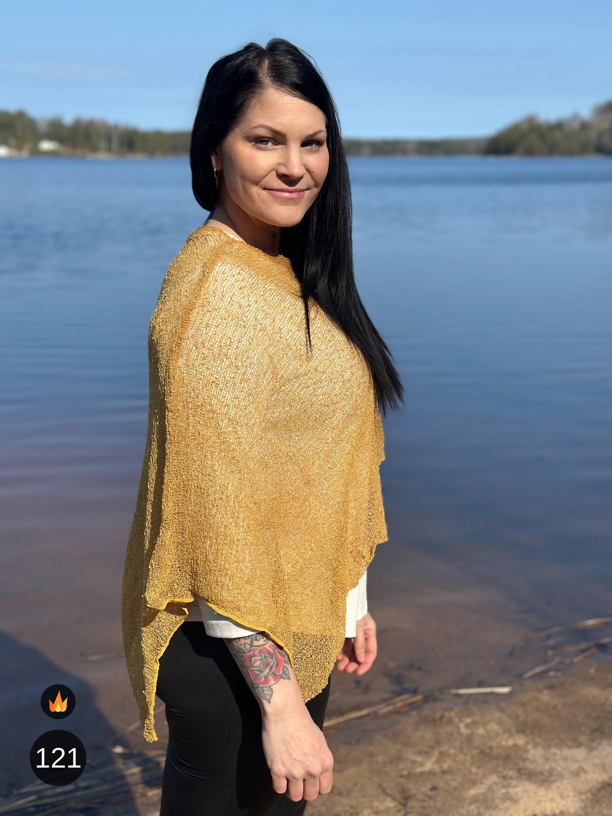 Kvinna bär Ella Cape i färg 121 Ljus senap, en varm färg i ljus senapsgul med gyllene skimmer, finstickad cape som draperas mjukt över axlarna, fotad vid en stilla sjö. Ulla Jacobsson Kläder.