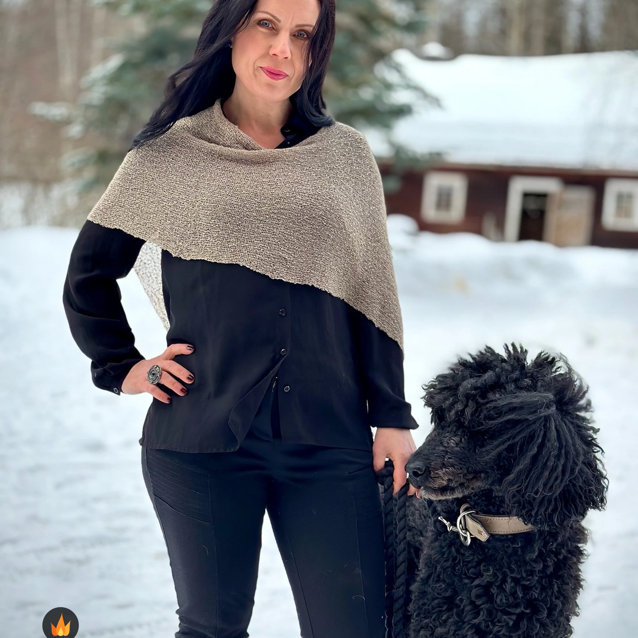 Kvinna bär Ella Cape i färg 123 Ljusbeige, en varm färg i ljusbeige med varm sandton, finstickad cape som draperas mjukt över axlarna, fotad i snön intill en svart lockig hund. Ulla Jacobsson Kläder.