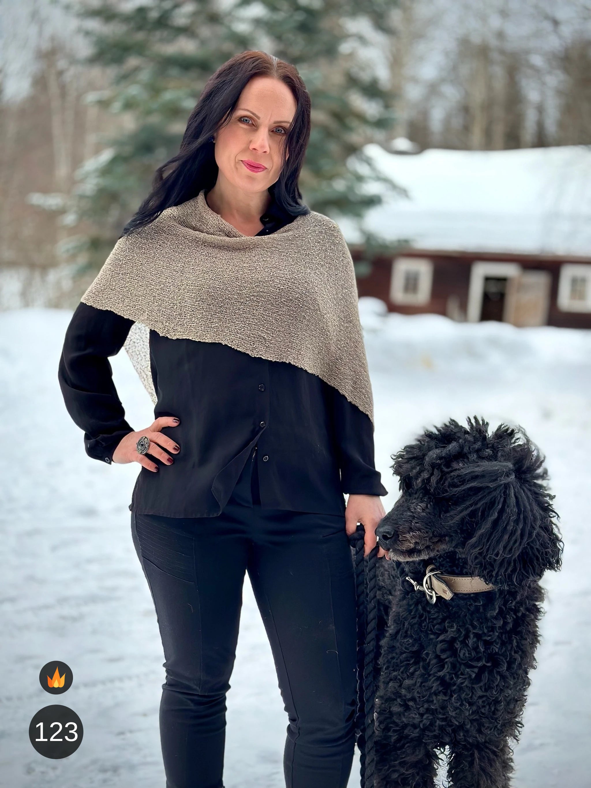 Kvinna bär Ella Cape i färg 123 Ljusbeige, en varm färg i ljusbeige med varm sandton, finstickad cape som draperas mjukt över axlarna, fotad i snön intill en svart lockig hund. Ulla Jacobsson Kläder.