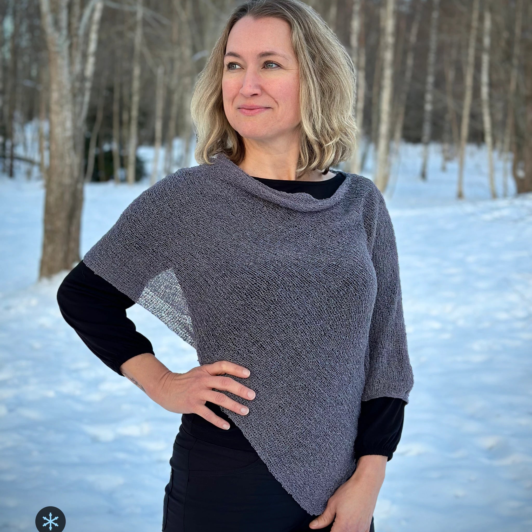 Kvinna bär Ella Cape i färg 124 Grålila, en kall färg i grålila med sval underton, finstickad cape som draperas mjukt över axlarna, fotad bland vinterns snöiga björkar. Ulla Jacobsson Kläder.
