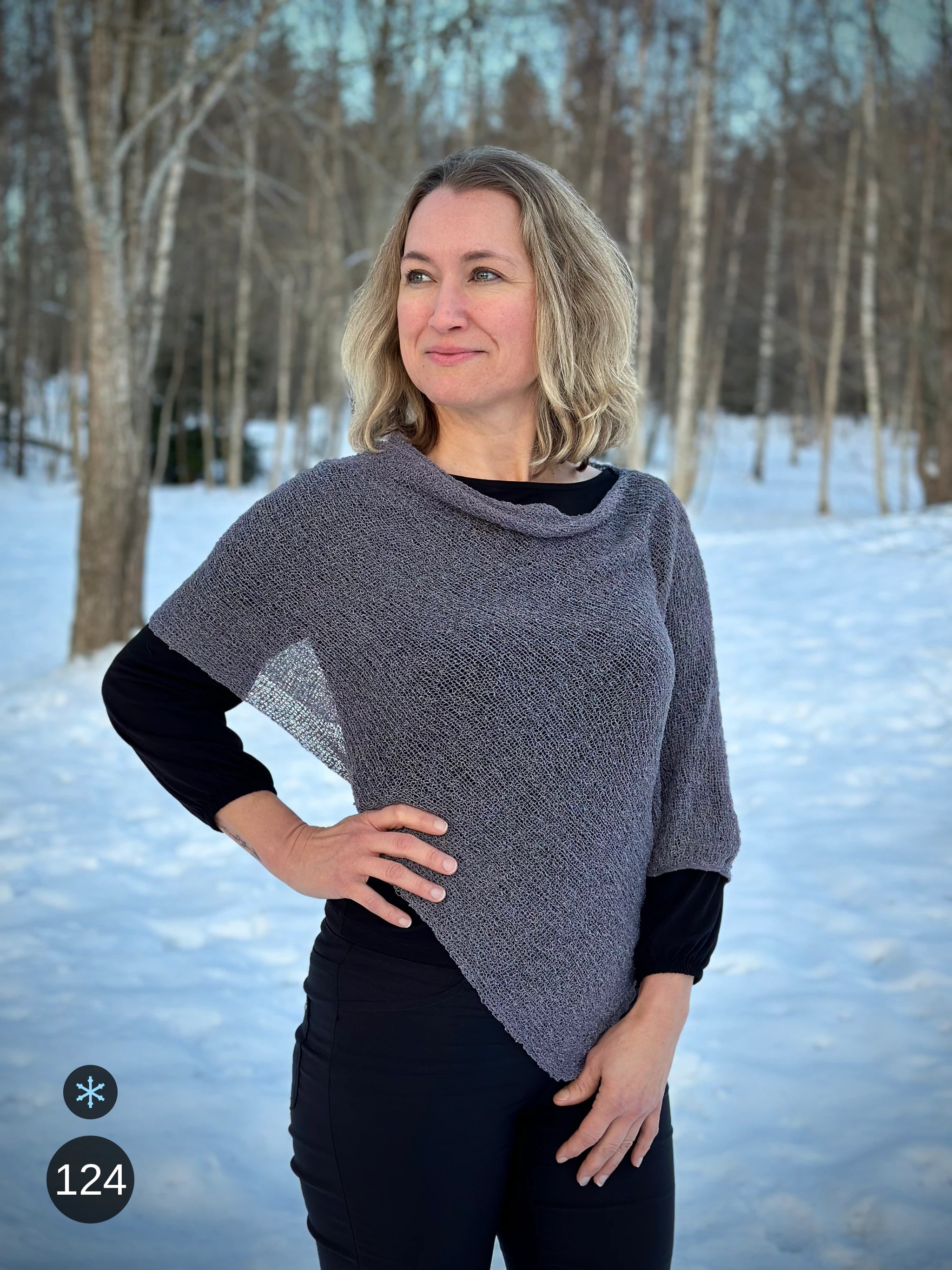 Kvinna bär Ella Cape i färg 124 Grålila, en kall färg i grålila med sval underton, finstickad cape som draperas mjukt över axlarna, fotad bland vinterns snöiga björkar. Ulla Jacobsson Kläder.