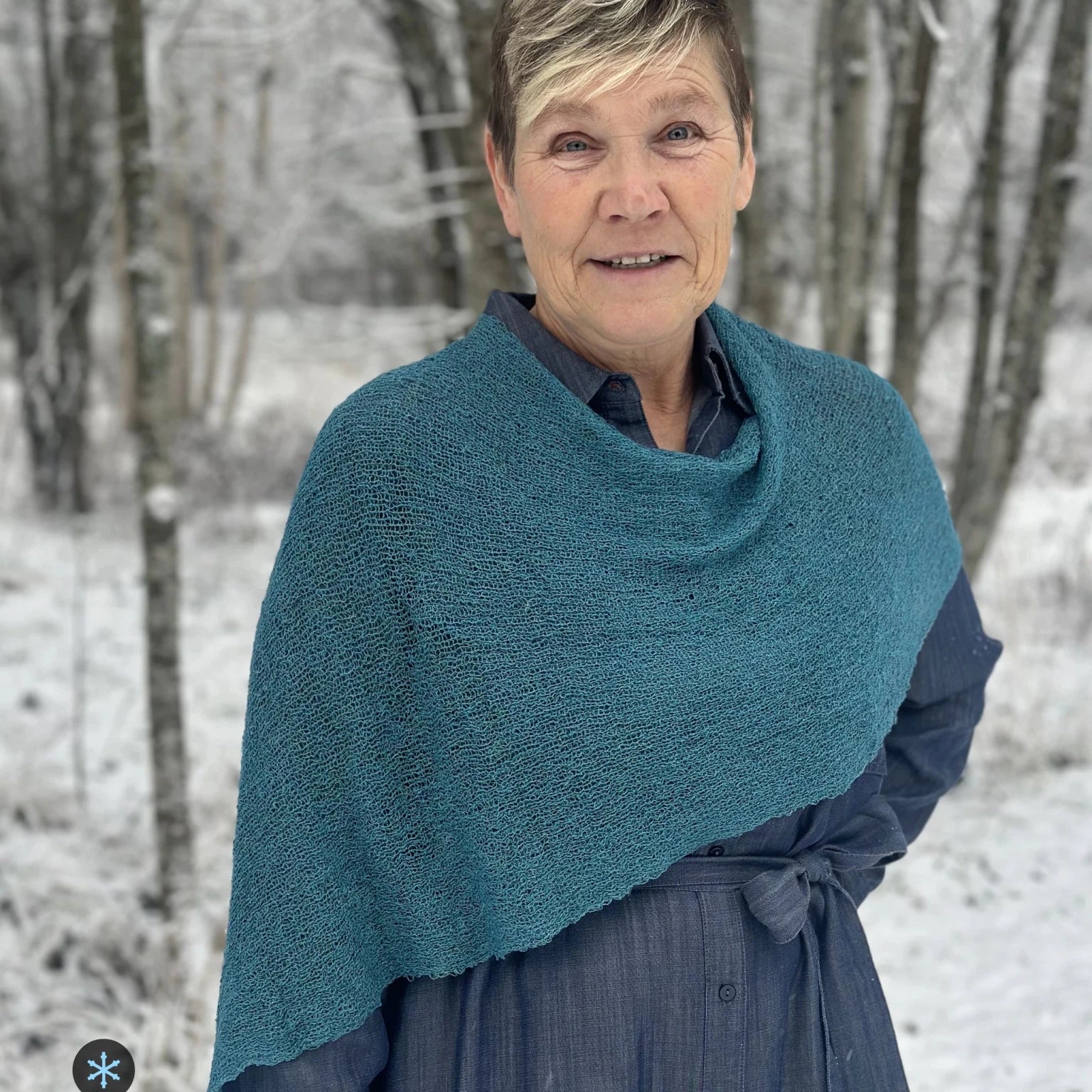Kvinna bär Ella Cape i färg 125 Mörk petrol, en kall färg, finstickad cape som draperas mjukt över axlarna, fotad bland snöiga björkar. Ulla Jacobsson Kläder.