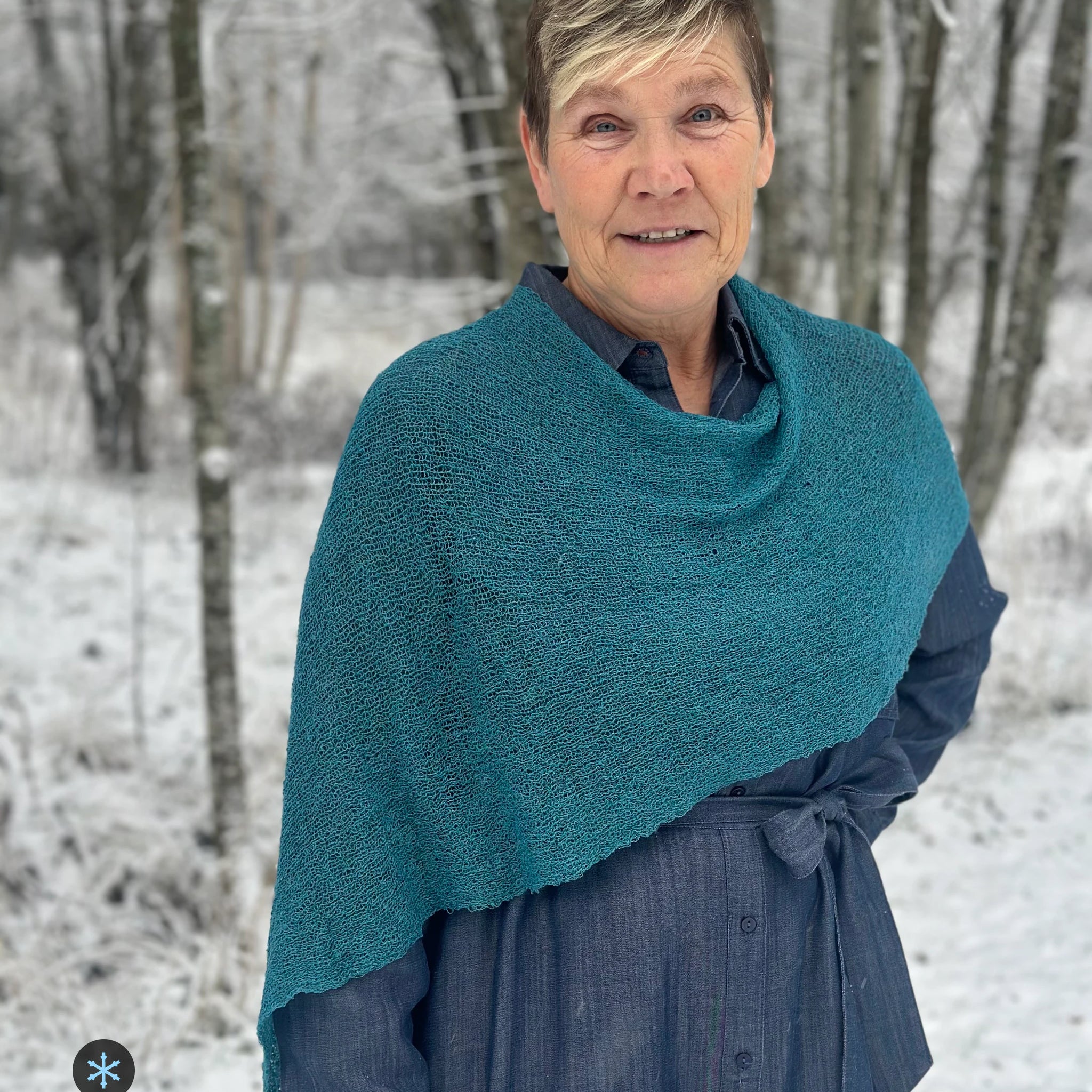 Kvinna bär Ella Cape i färg 125 Mörk petrol, en kall färg, finstickad cape som draperas mjukt över axlarna, fotad bland snöiga björkar. Ulla Jacobsson Kläder.