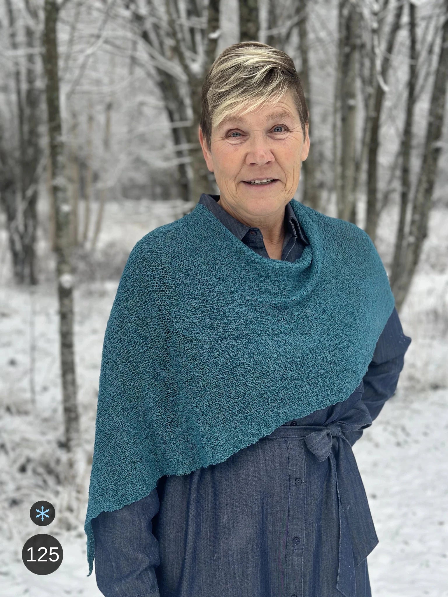 Kvinna bär Ella Cape i färg 125 Mörk petrol, en kall färg, finstickad cape som draperas mjukt över axlarna, fotad bland snöiga björkar. Ulla Jacobsson Kläder.