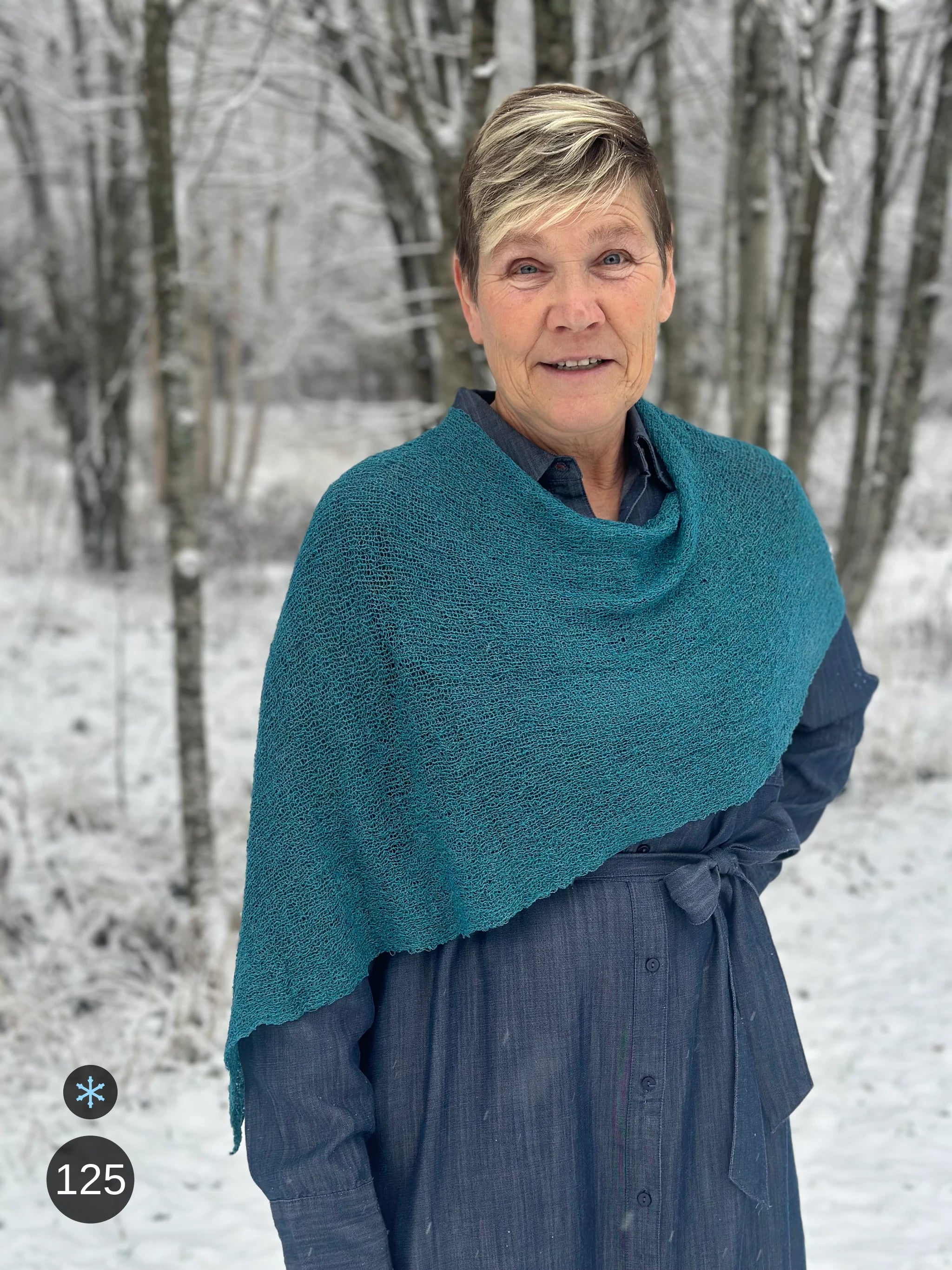 Kvinna bär Ella Cape i färg 125 Mörk petrol, en kall färg, finstickad cape som draperas mjukt över axlarna, fotad bland snöiga björkar. Ulla Jacobsson Kläder.