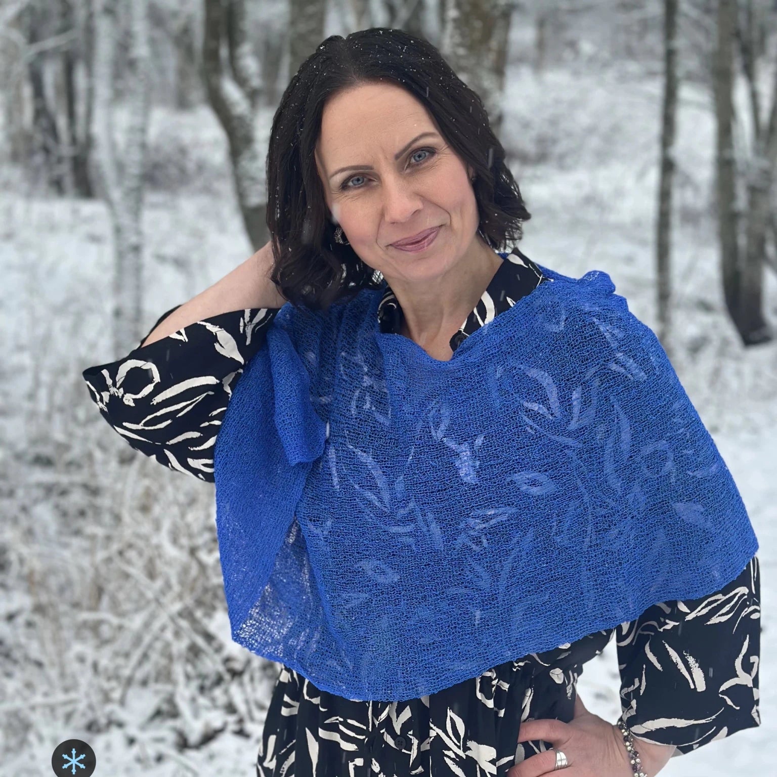 Kvinna bär Ella Cape i färg 126 Sverigeblå, en kall färg, finstickad cape som draperas mjukt över axlarna, fotad bland vinterträd. Ulla Jacobsson Kläder.