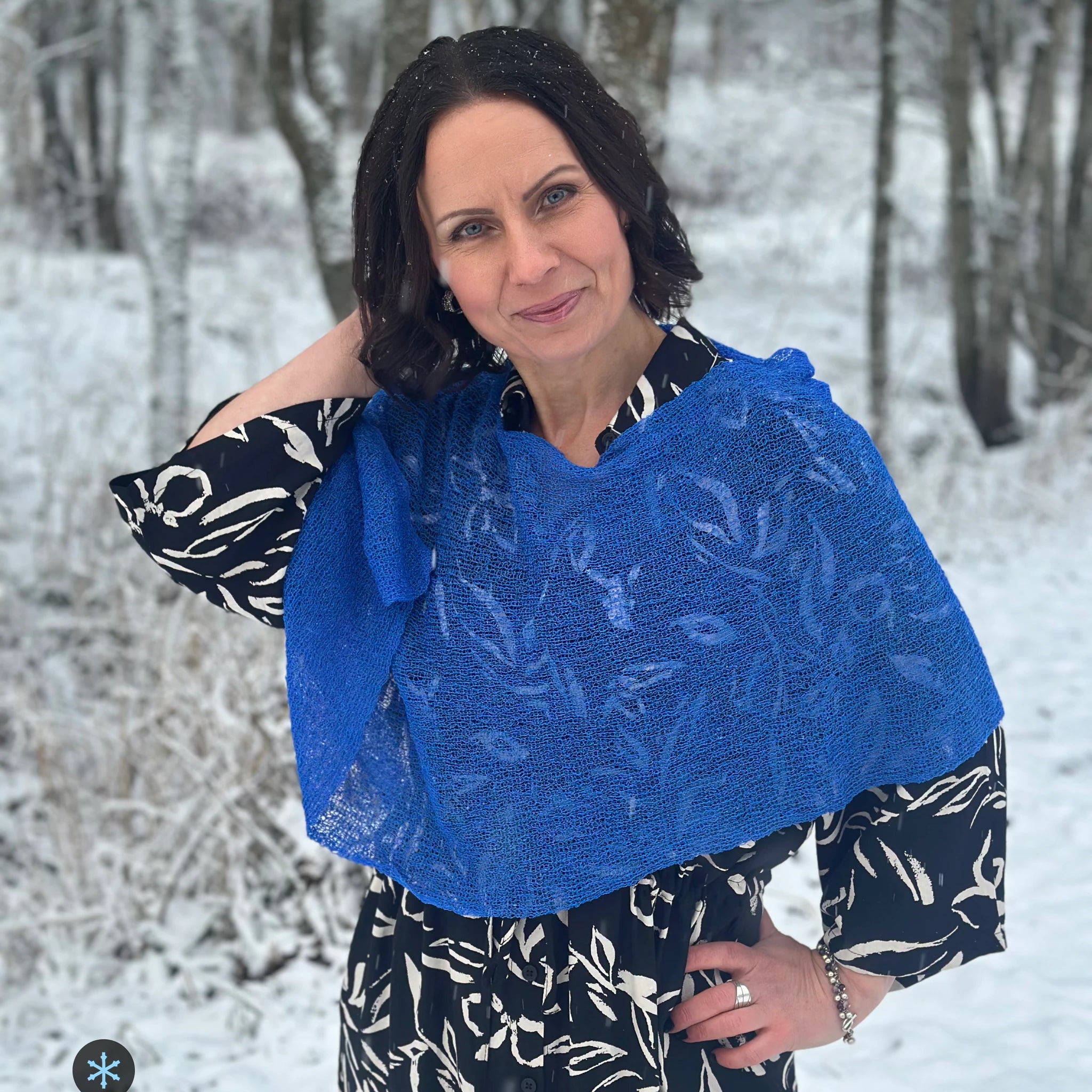 Kvinna bär Ella Cape i färg 126 Sverigeblå, en kall färg, finstickad cape som draperas mjukt över axlarna, fotad bland vinterträd. Ulla Jacobsson Kläder.
