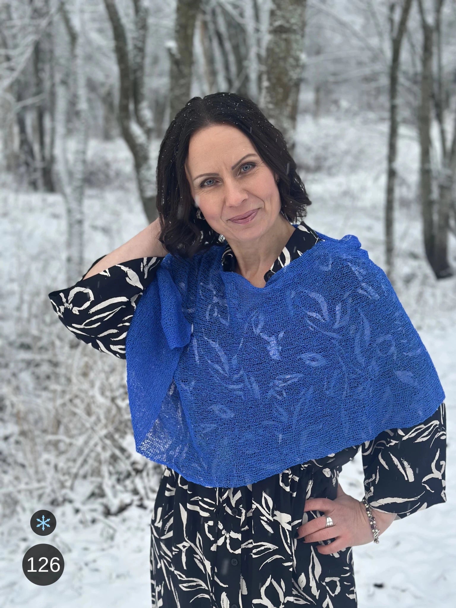 Kvinna bär Ella Cape i färg 126 Sverigeblå, en kall färg, finstickad cape som draperas mjukt över axlarna, fotad bland vinterträd. Ulla Jacobsson Kläder.