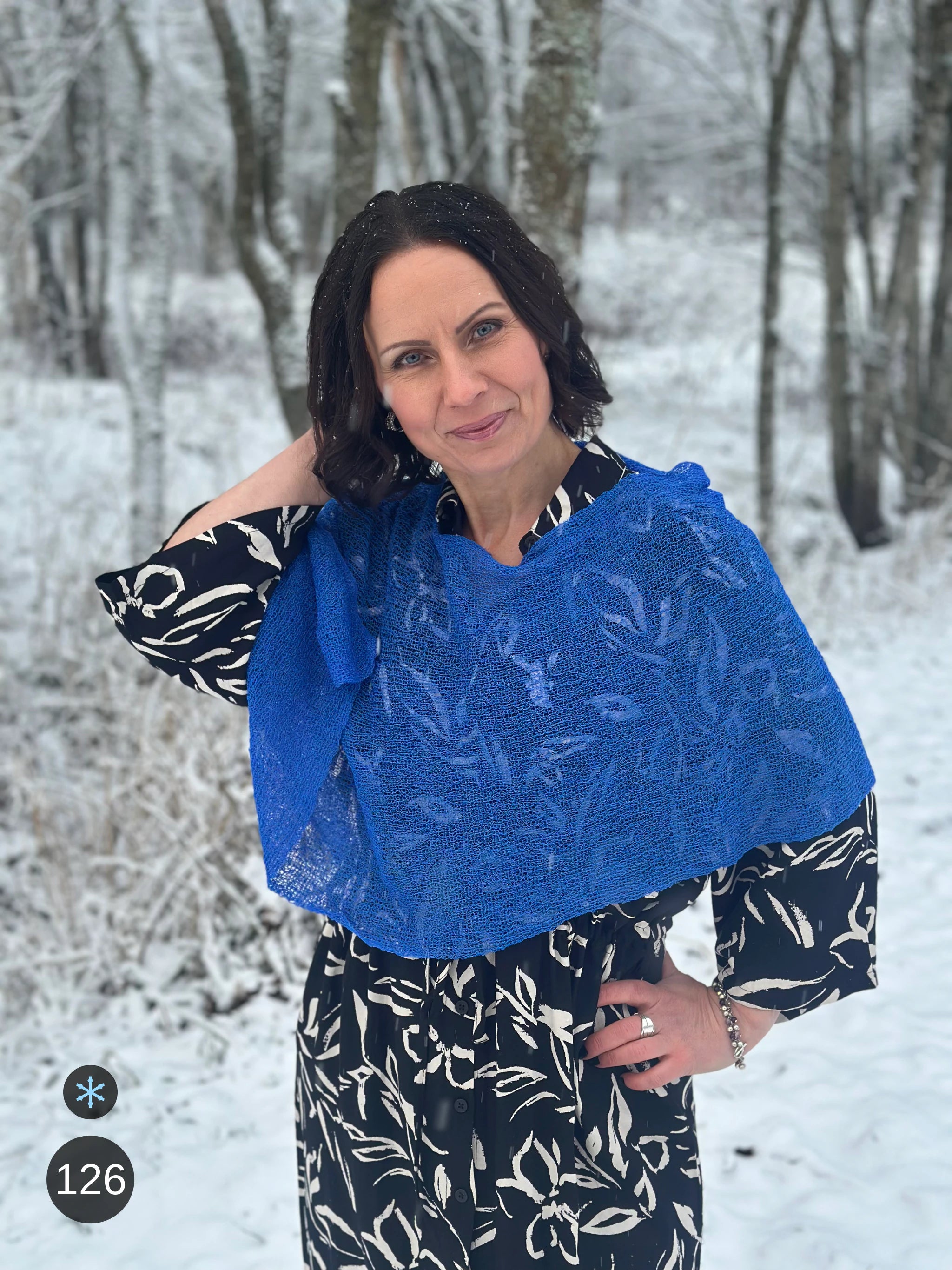 Kvinna bär Ella Cape i färg 126 Sverigeblå, en kall färg, finstickad cape som draperas mjukt över axlarna, fotad bland vinterträd. Ulla Jacobsson Kläder.