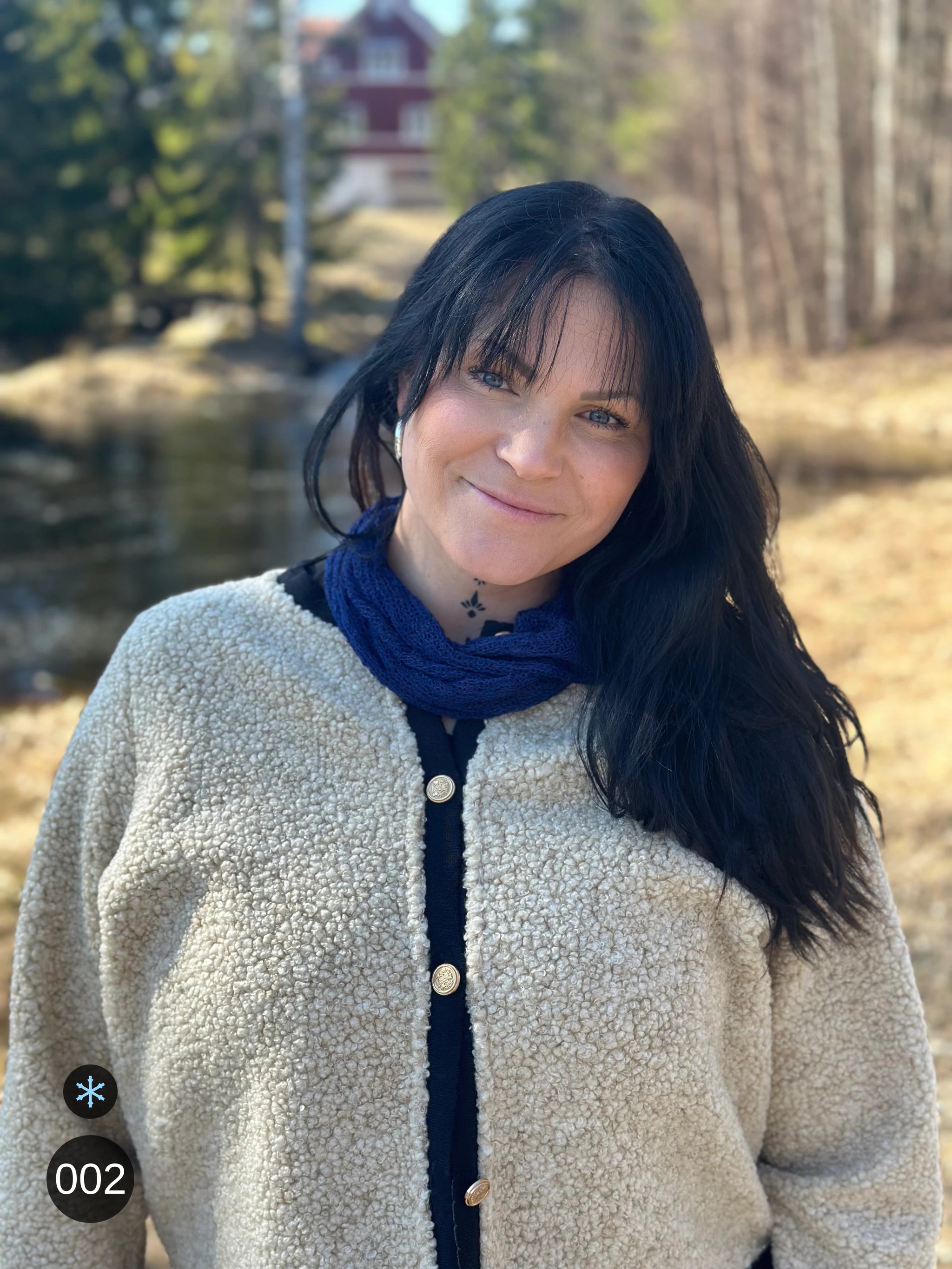 Kvinna bär Eva Tubsjal i färg 002 Marinblå, en kall färg i djup marinblå, liten finstickad tubsjal buren mjukt runt halsen, fotad framför ett rött hus i vårvinterljus. Ulla Jacobsson Kläder.