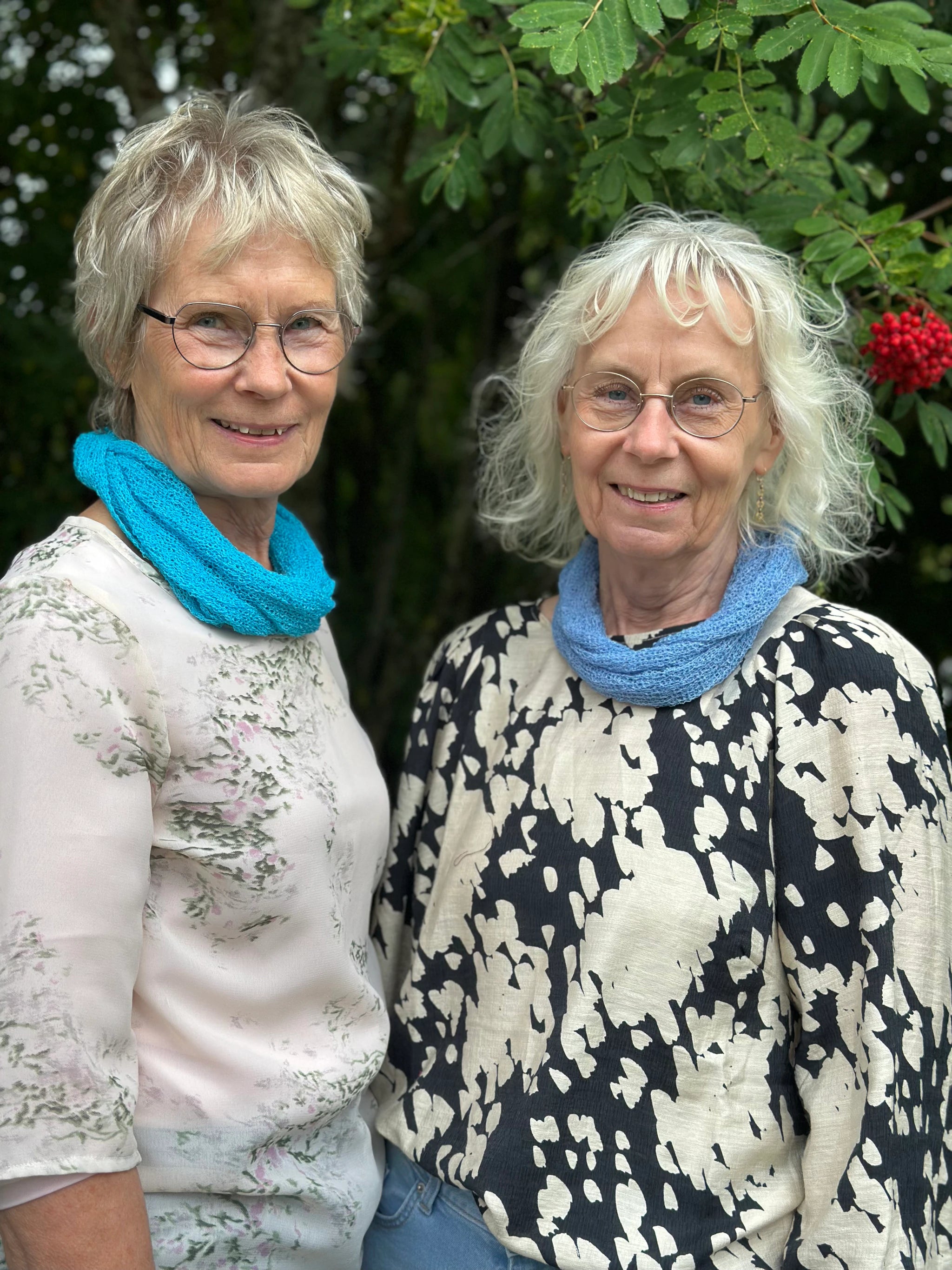 Två kvinnor bär Eva Tubsjal, den ena i 004 Turkos och den andra i 033 Ljusblå. Små finstickade tubsjalar burna mjukt runt halsen, fotade under en rönn med röda bär. Ulla Jacobsson Kläder.