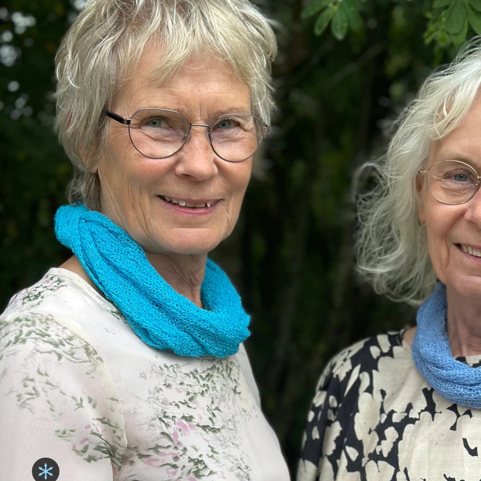 Kvinna bär Eva Tubsjal i färg 004 Turkos, en kall färg i klar, frisk turkos, liten finstickad tubsjal buren mjukt runt halsen, fotad utomhus i grönska. Ulla Jacobsson Kläder.