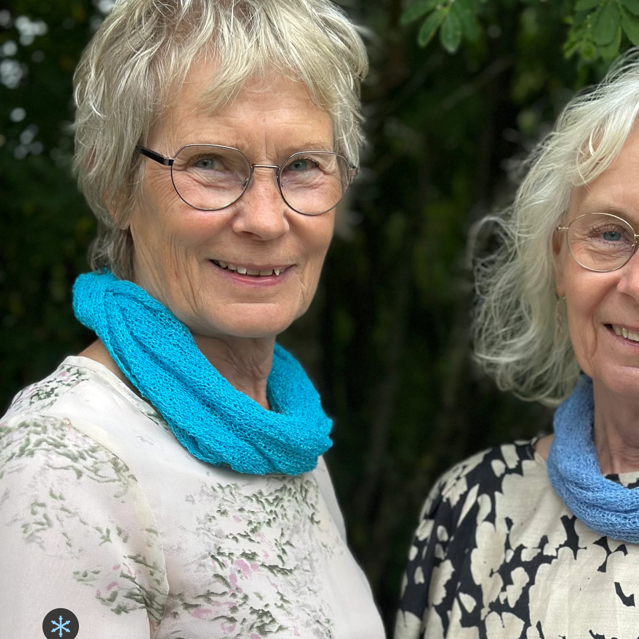 Kvinna bär Eva Tubsjal i färg 004 Turkos, en kall färg i klar, frisk turkos, liten finstickad tubsjal buren mjukt runt halsen, fotad utomhus i grönska. Ulla Jacobsson Kläder.