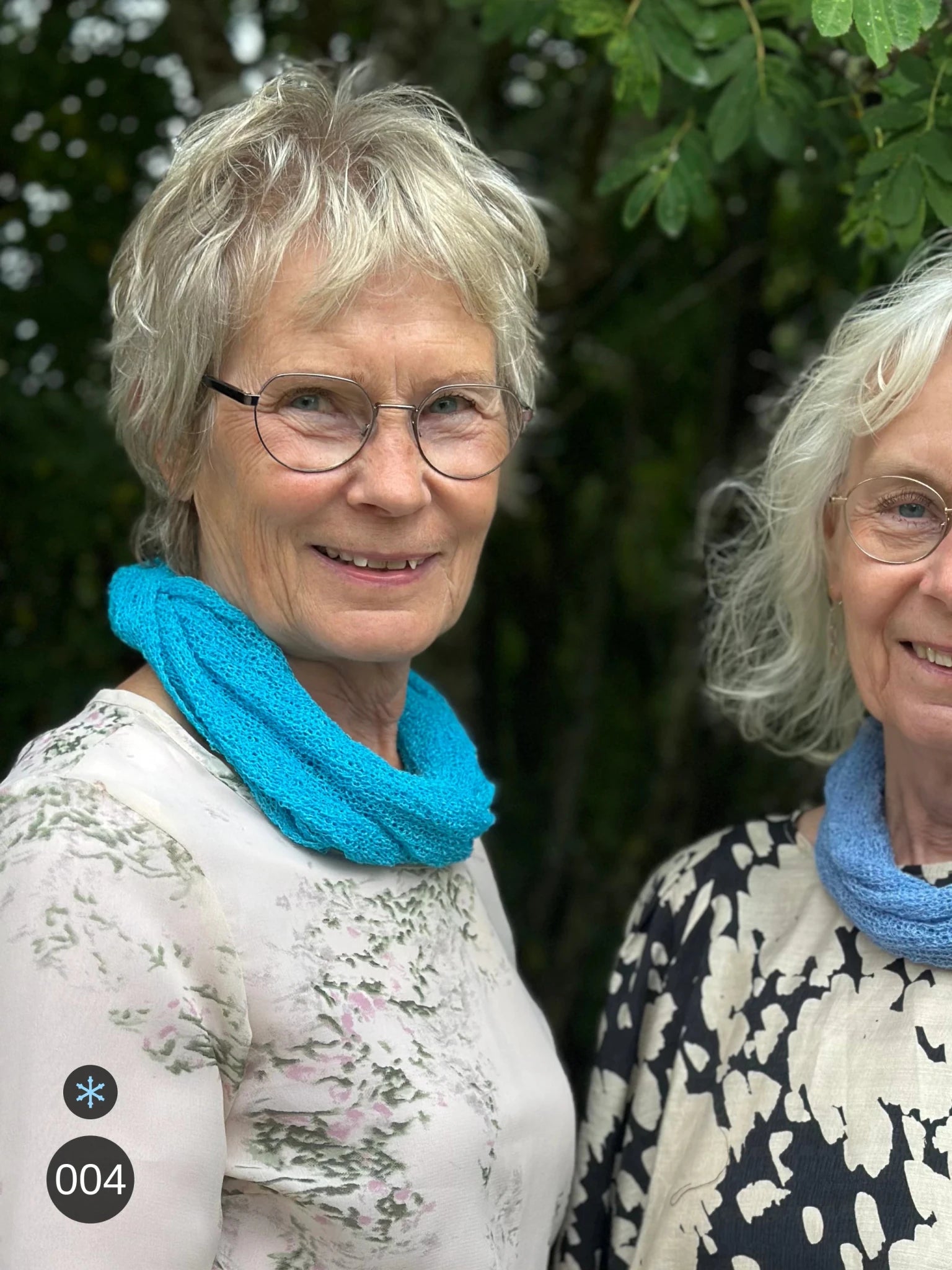 Kvinna bär Eva Tubsjal i färg 004 Turkos, en kall färg i klar, frisk turkos, liten finstickad tubsjal buren mjukt runt halsen, fotad utomhus i grönska. Ulla Jacobsson Kläder.