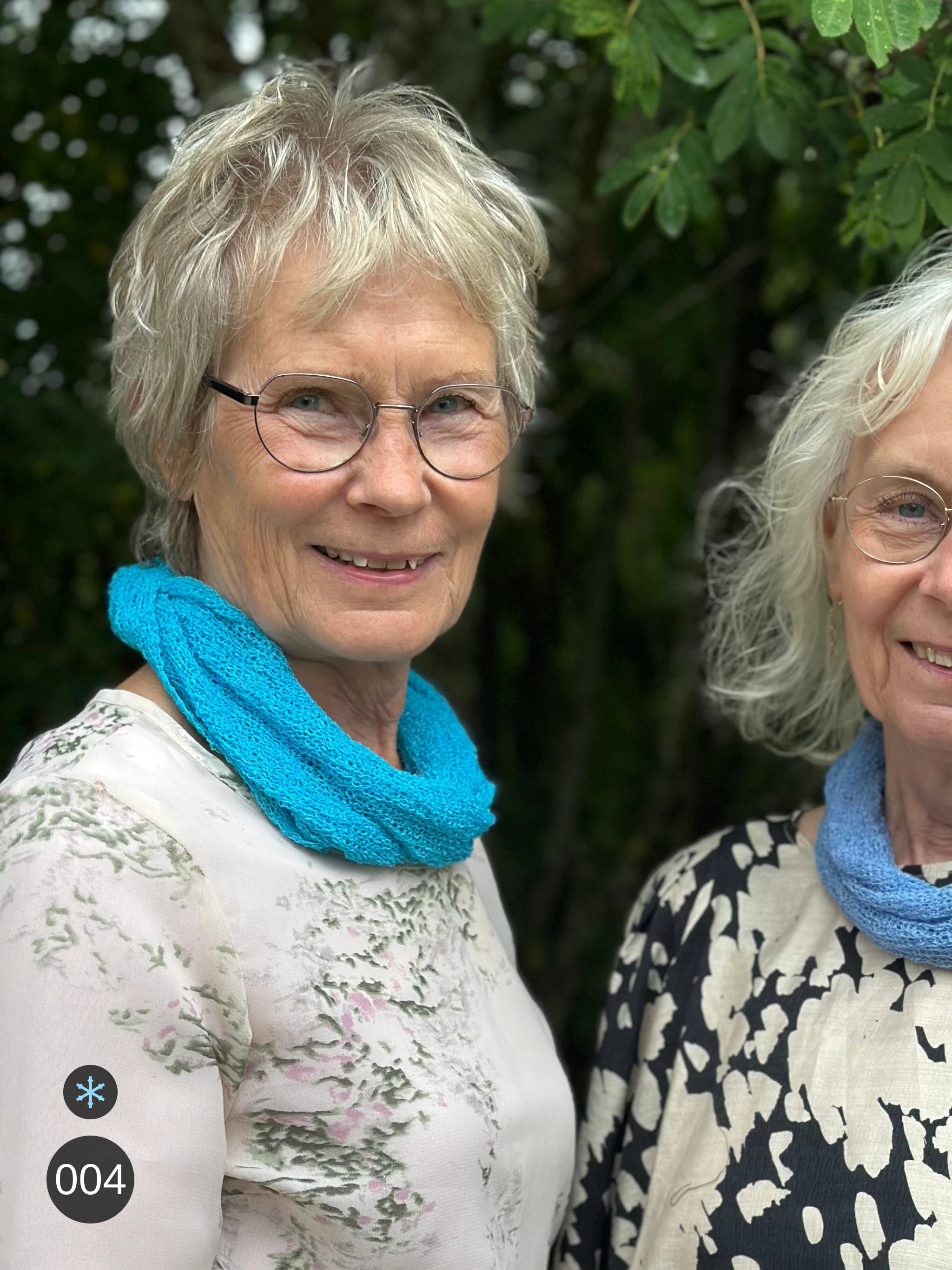 Kvinna bär Eva Tubsjal i färg 004 Turkos, en kall färg i klar, frisk turkos, liten finstickad tubsjal buren mjukt runt halsen, fotad utomhus i grönska. Ulla Jacobsson Kläder.