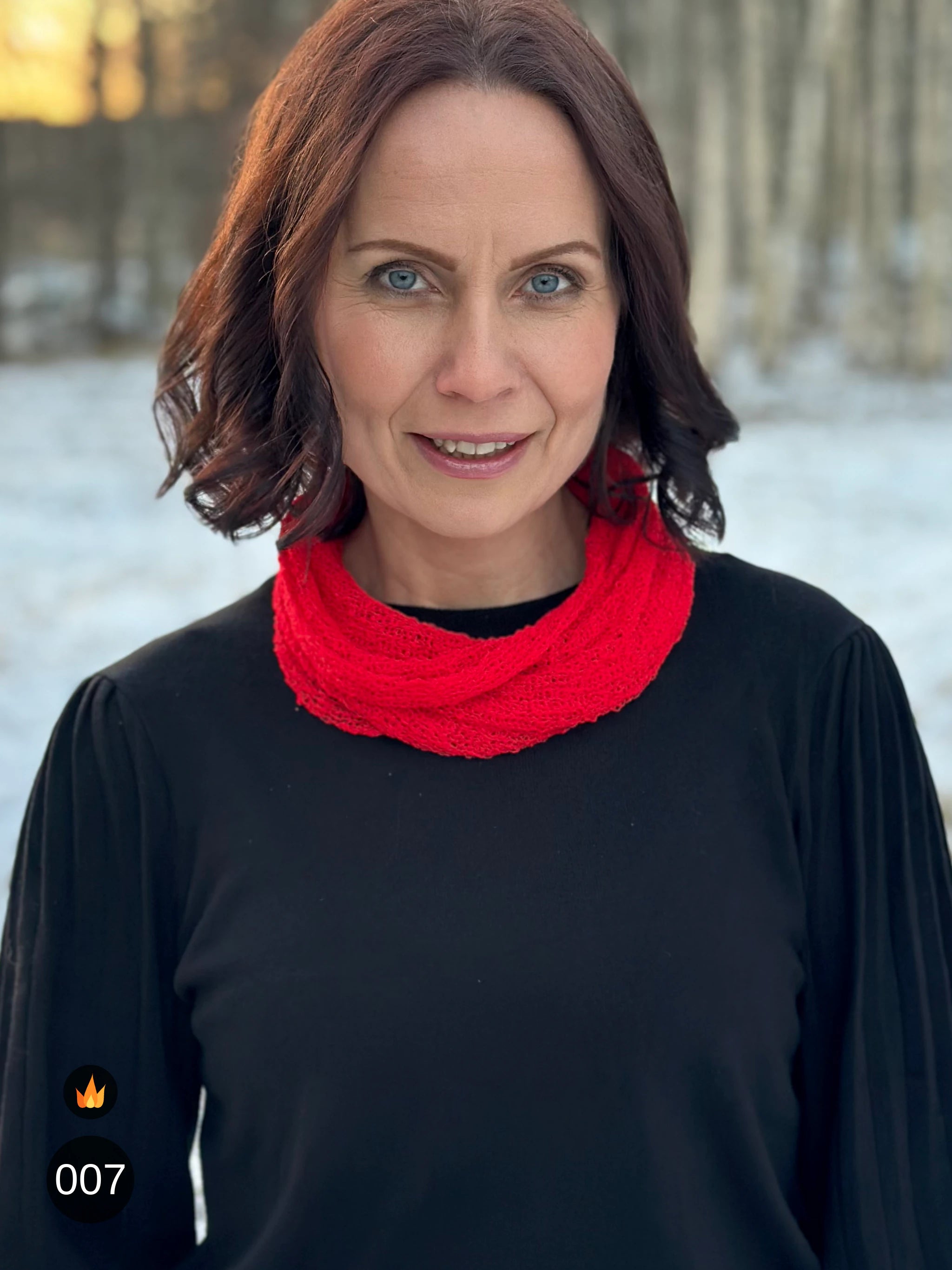 Kvinna bär Eva Tubsjal i färg 007 Klarröd, en varm färg i klar signalröd med lätt orange glöd, liten finstickad tubsjal buren mjukt runt halsen, fotad i snöig björkskog. Ulla Jacobsson Kläder.