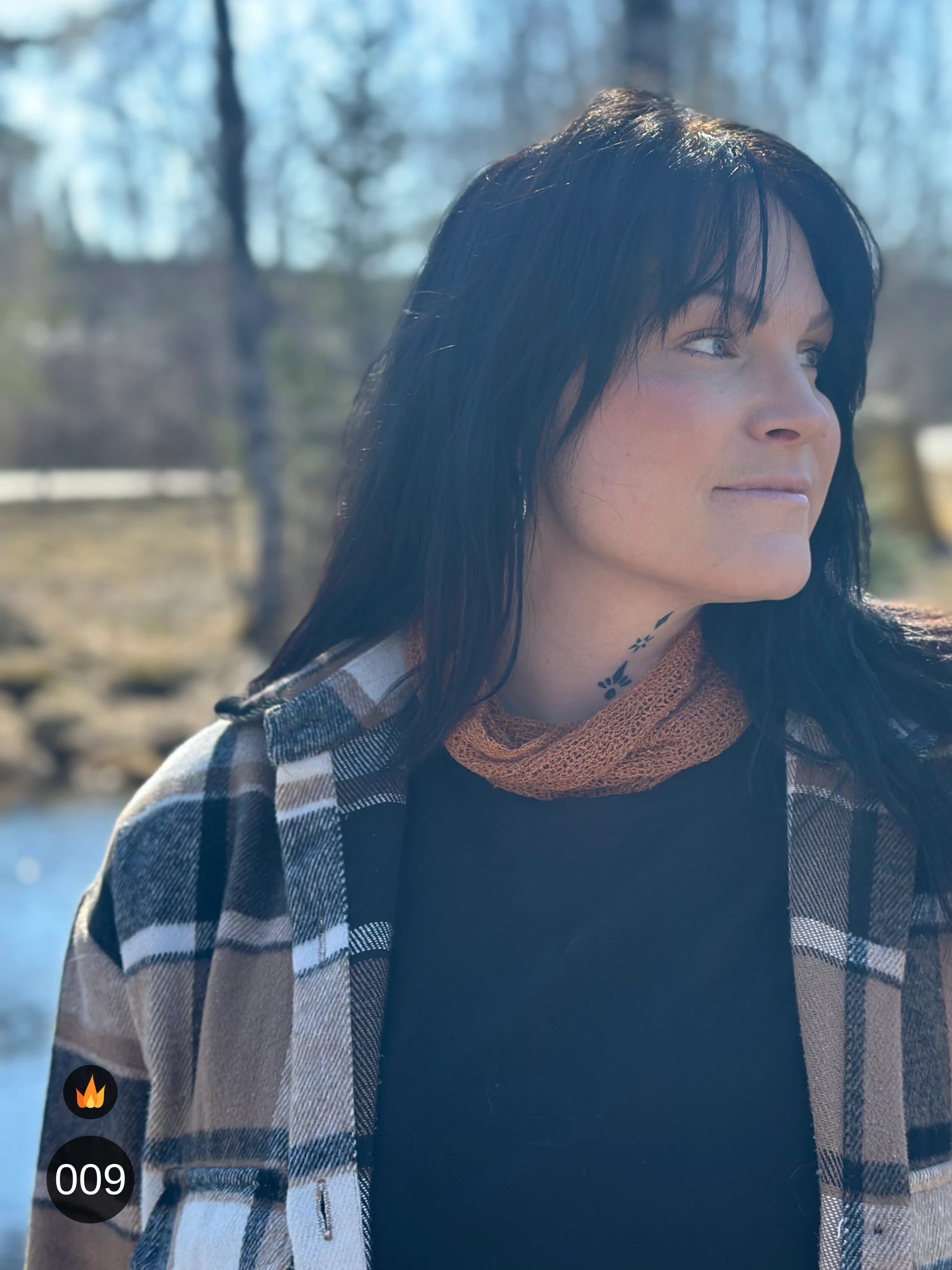 Kvinna bär Eva Tubsjal i färg 009 Mocka, en varm färg i varm mockabrun, liten finstickad tubsjal buren mjukt runt halsen, fotad vid vattnet en tidig vårdag. Ulla Jacobsson Kläder.