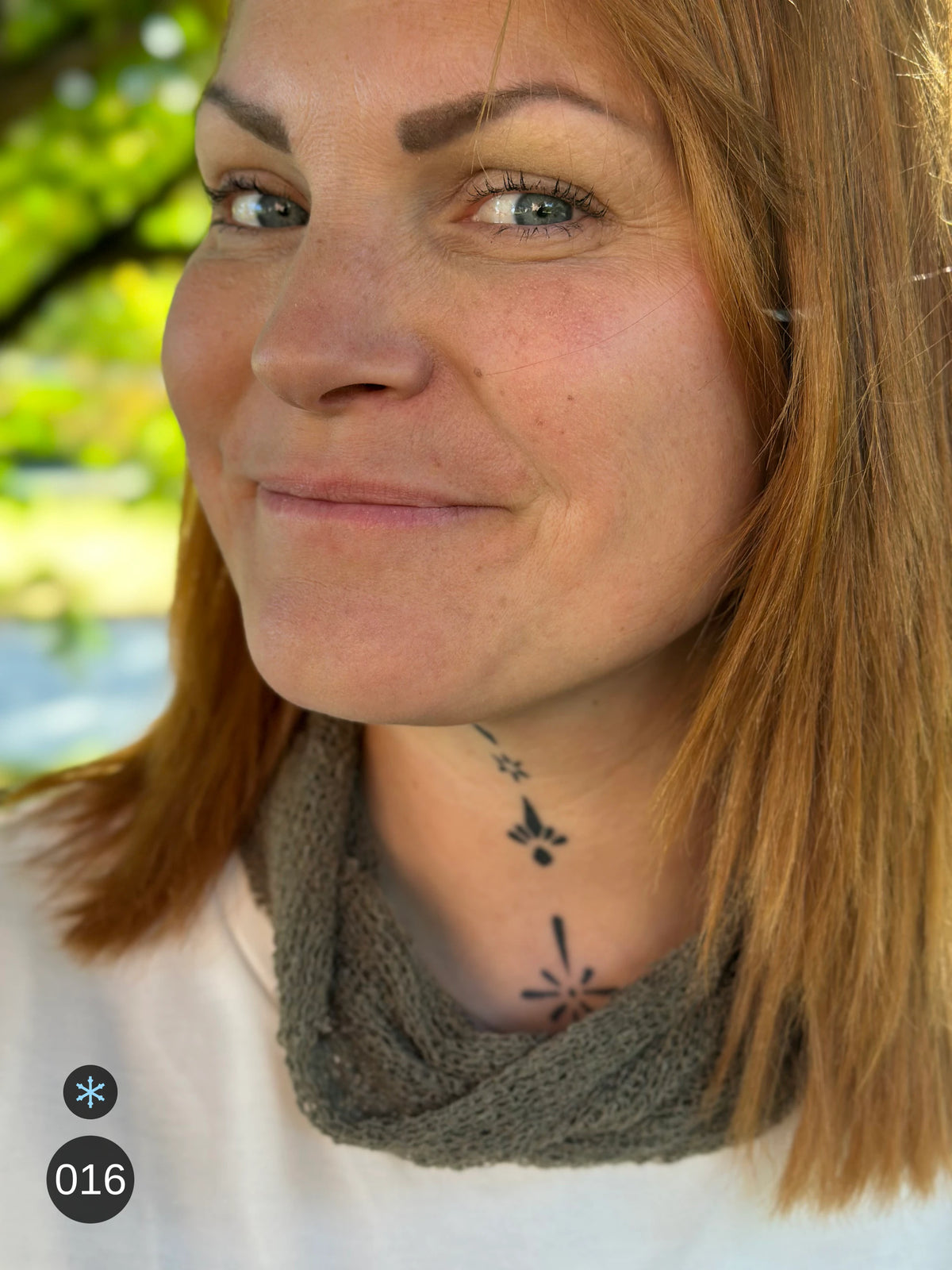 Kvinna bär Eva Tubsjal i färg 016 Lavgrön, en kall färg i dämpad gråbrun grönton som lav på sten, liten finstickad tubsjal buren som krage, fotad i höstsol under en lönn. Ulla Jacobsson Kläder.