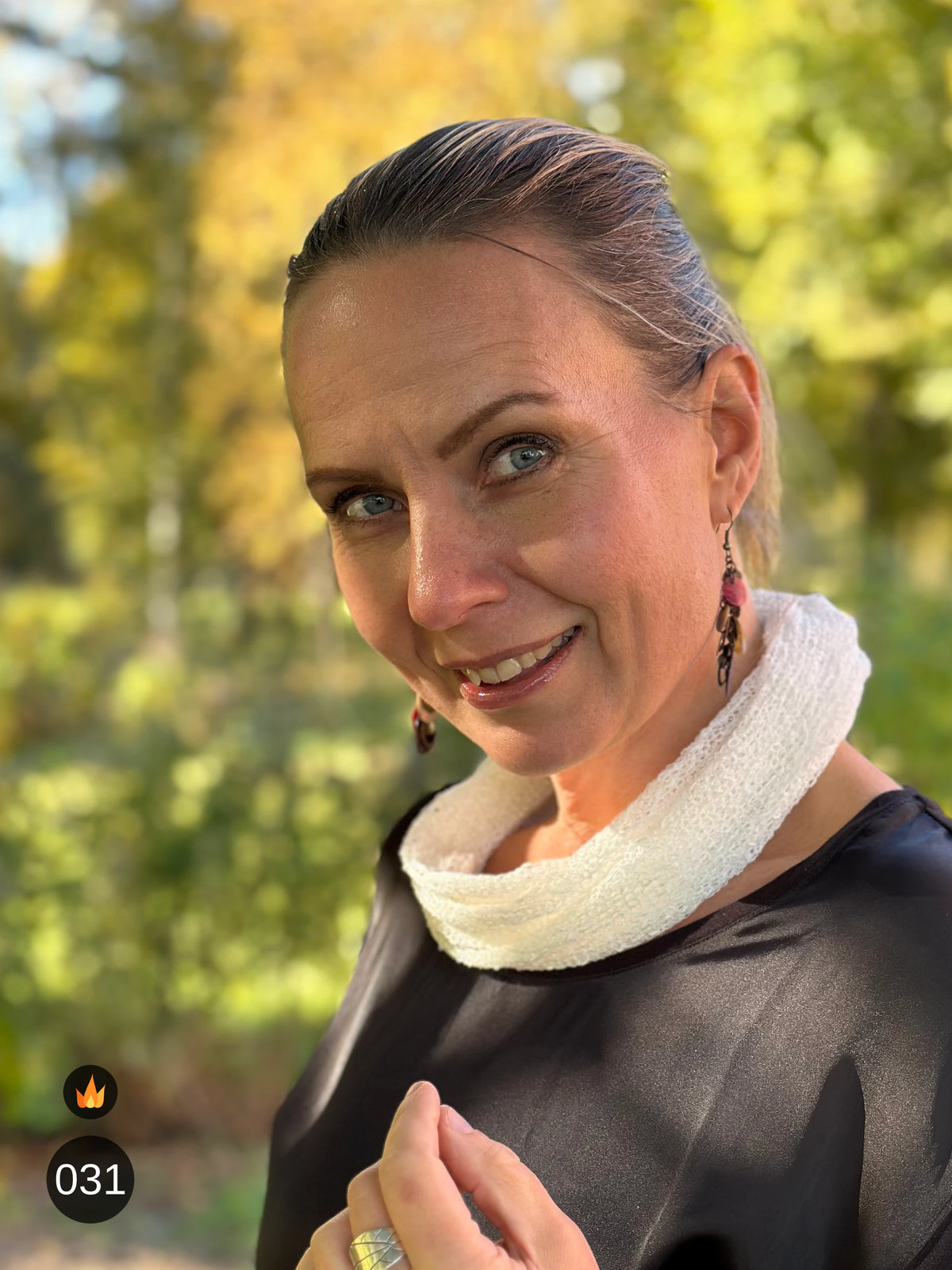 Kvinna bär Eva Tubsjal i färg 031 Offwhite, en varm färg i mjuk naturvit med gräddig underton, liten finstickad tubsjal buren som krage, fotad i höstsol under en lönn. Ulla Jacobsson Kläder.