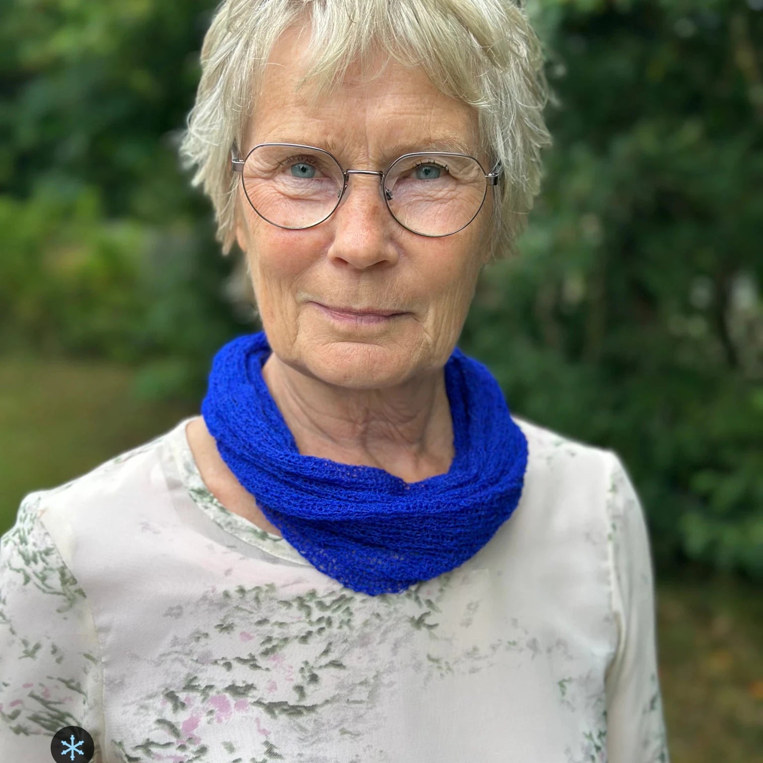 Kvinna bär Eva Tubsjal i färg 032 Klarblå, en kall färg i intensiv klarblå kobolt, liten finstickad tubsjal buren mjukt runt halsen, fotad utomhus i grönska. Ulla Jacobsson Kläder.