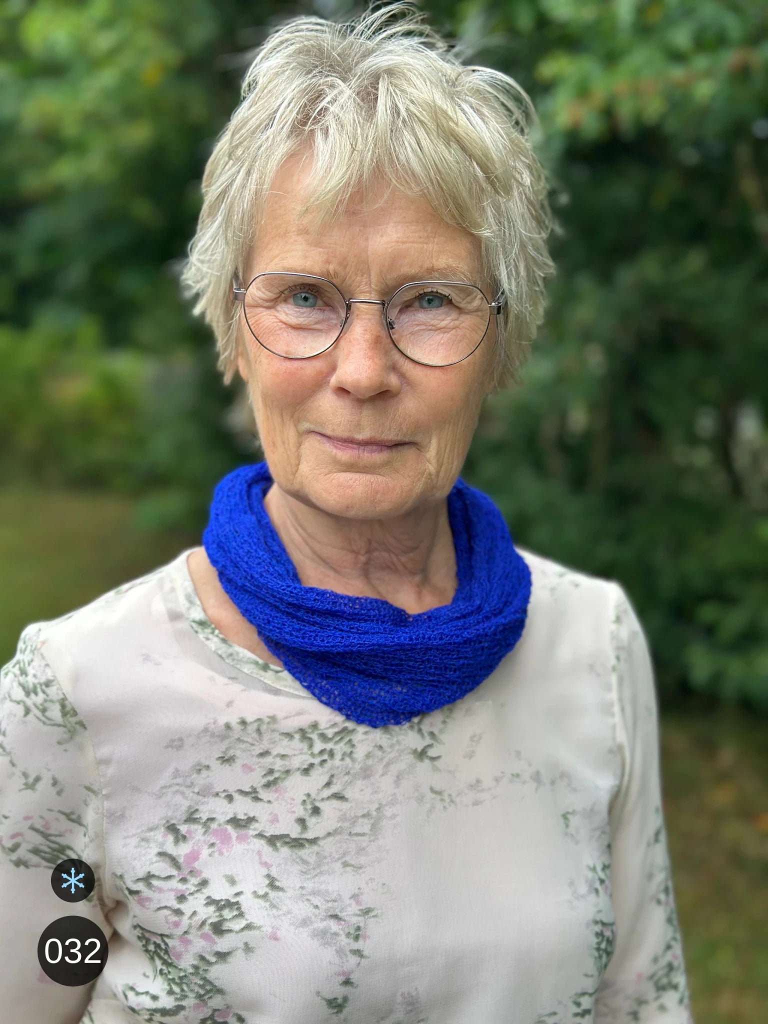 Kvinna bär Eva Tubsjal i färg 032 Klarblå, en kall färg i intensiv klarblå kobolt, liten finstickad tubsjal buren mjukt runt halsen, fotad utomhus i grönska. Ulla Jacobsson Kläder.