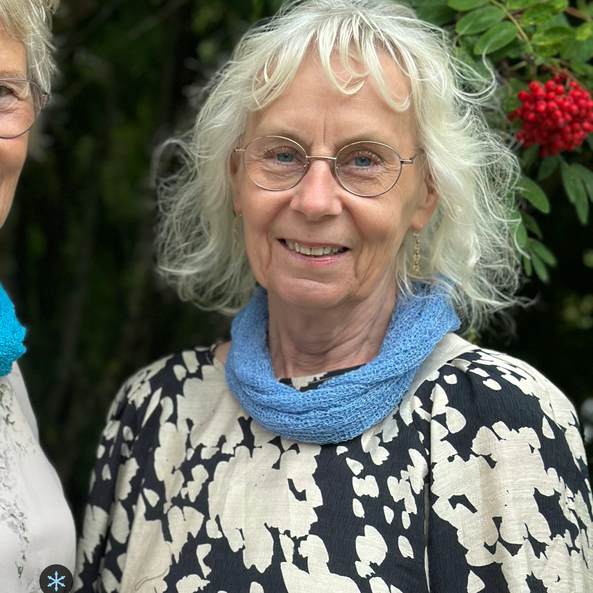 Kvinna bär Eva Tubsjal i färg 033 Ljusblå, en kall färg i dämpad ljusblå, liten finstickad tubsjal buren mjukt runt halsen, fotad utomhus i grönska. Ulla Jacobsson Kläder.