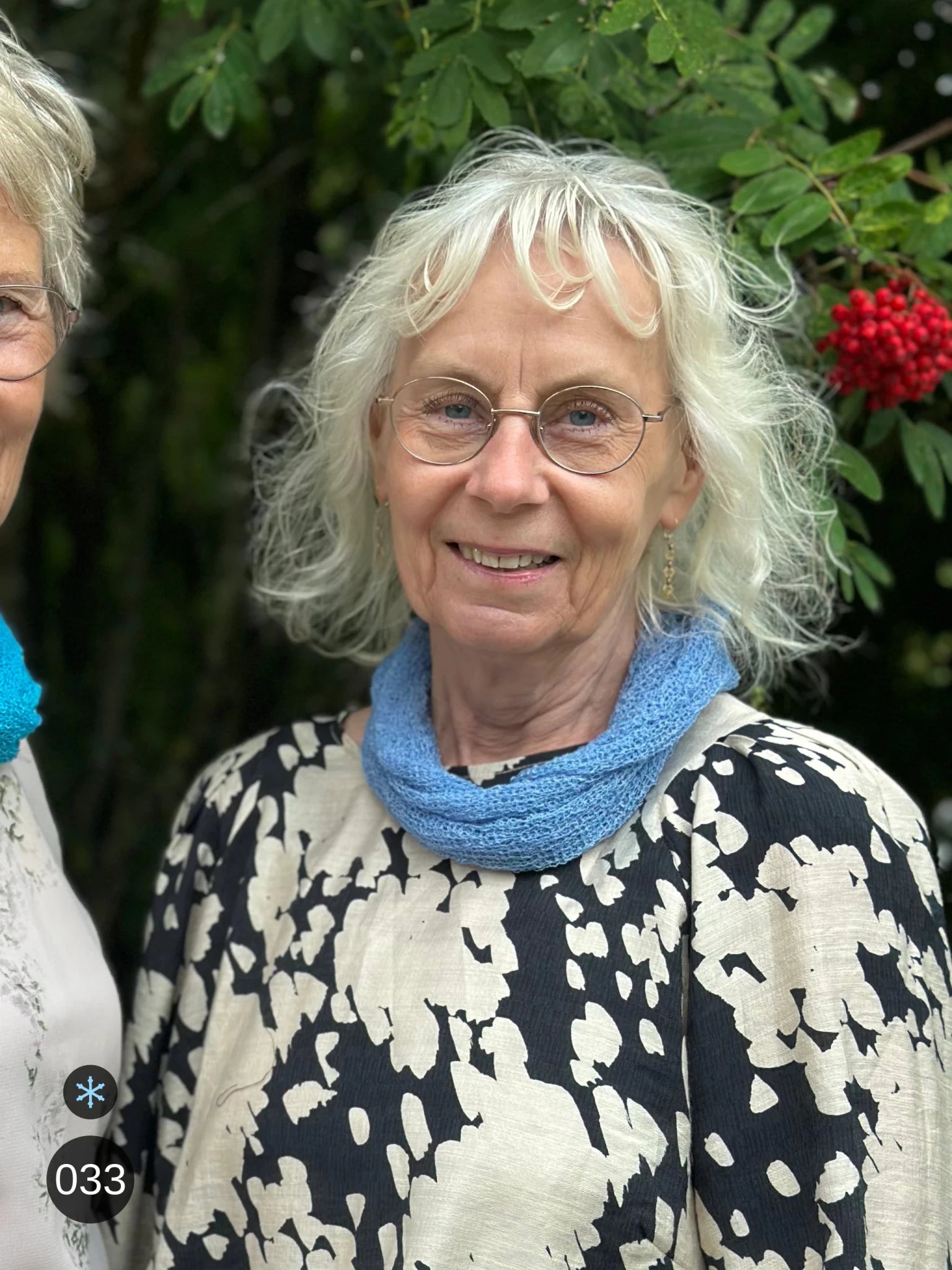 Kvinna bär Eva Tubsjal i färg 033 Ljusblå, en kall färg i dämpad ljusblå, liten finstickad tubsjal buren mjukt runt halsen, fotad utomhus i grönska. Ulla Jacobsson Kläder.